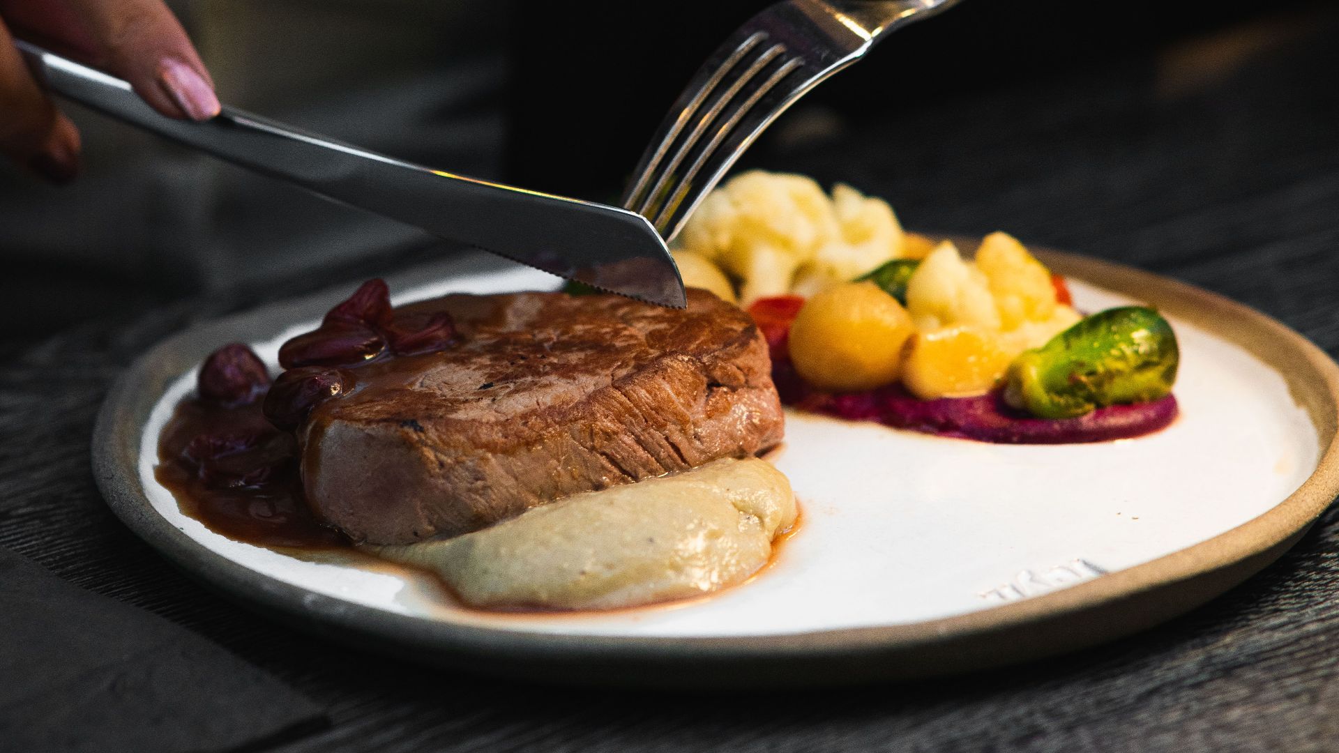 A close-up of a gourmet steak meal with vegetables, elegantly plated, showing hands using fork and knife.