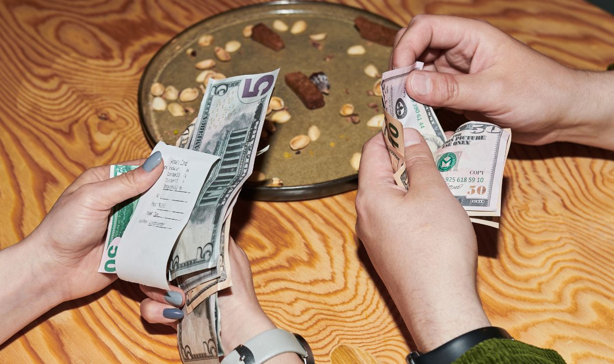 Close up of young adults splitting restaurant bill, counting cash and checking receipt at table. Showing shared expenses, budgeting, dining costs, consumer finance content