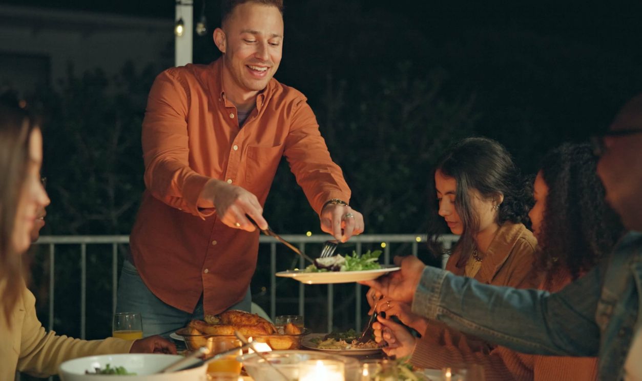 Friends, night and man with knife for chicken on patio, happy or talk for food at new years eve party. Women, group and cutting at table for chat, memory or giving plate for meat, diversity or event