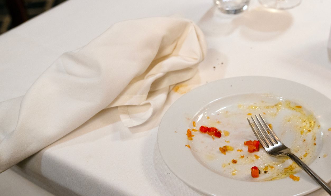 A close-up view of a messy finished dinner table with a dirty plate, used fork, crumpled napkin, and wine glasses after a meal.