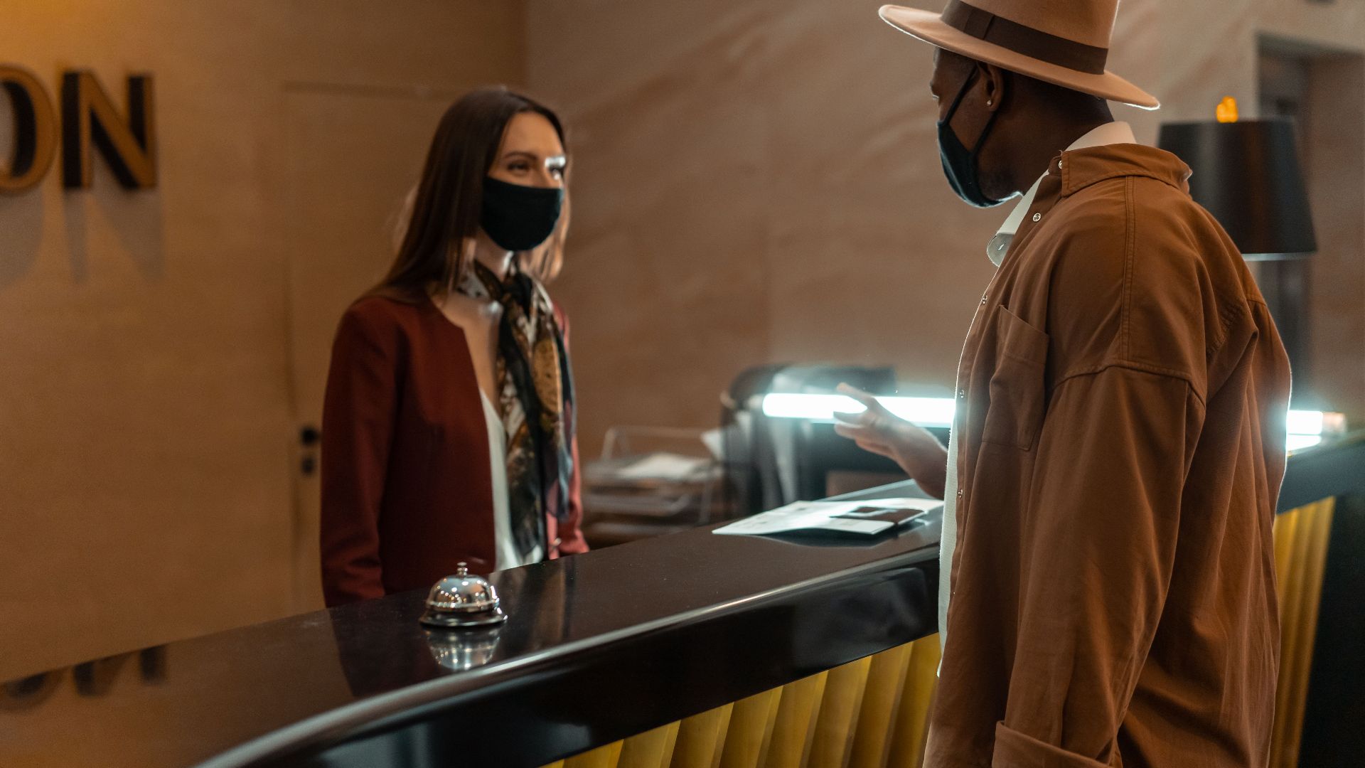 A man at a hotel reception talking to a staff member. Both wear face masks.