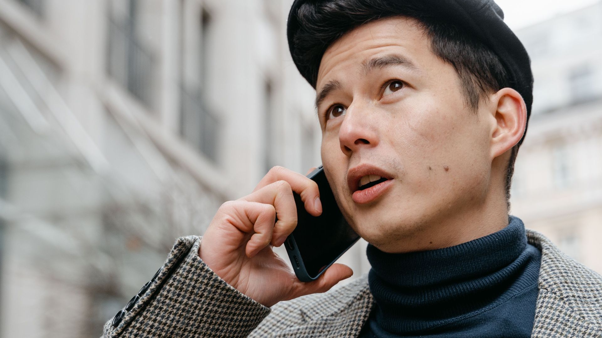Stylish man in a city talking on a smartphone outdoors, wearing a coat and hat.
