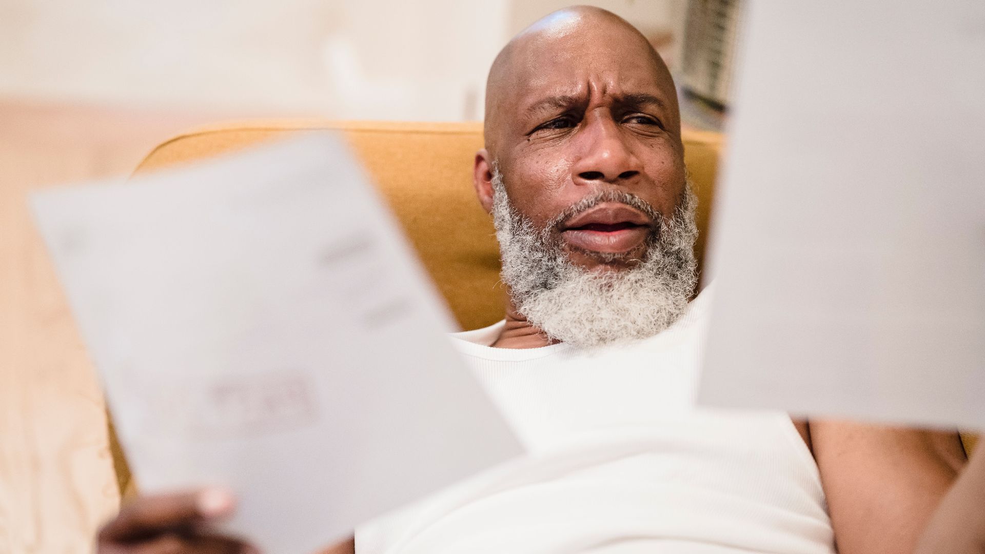 A middle-aged man looks concerned as he reviews household bills, sitting in a comfortable chair indoors.