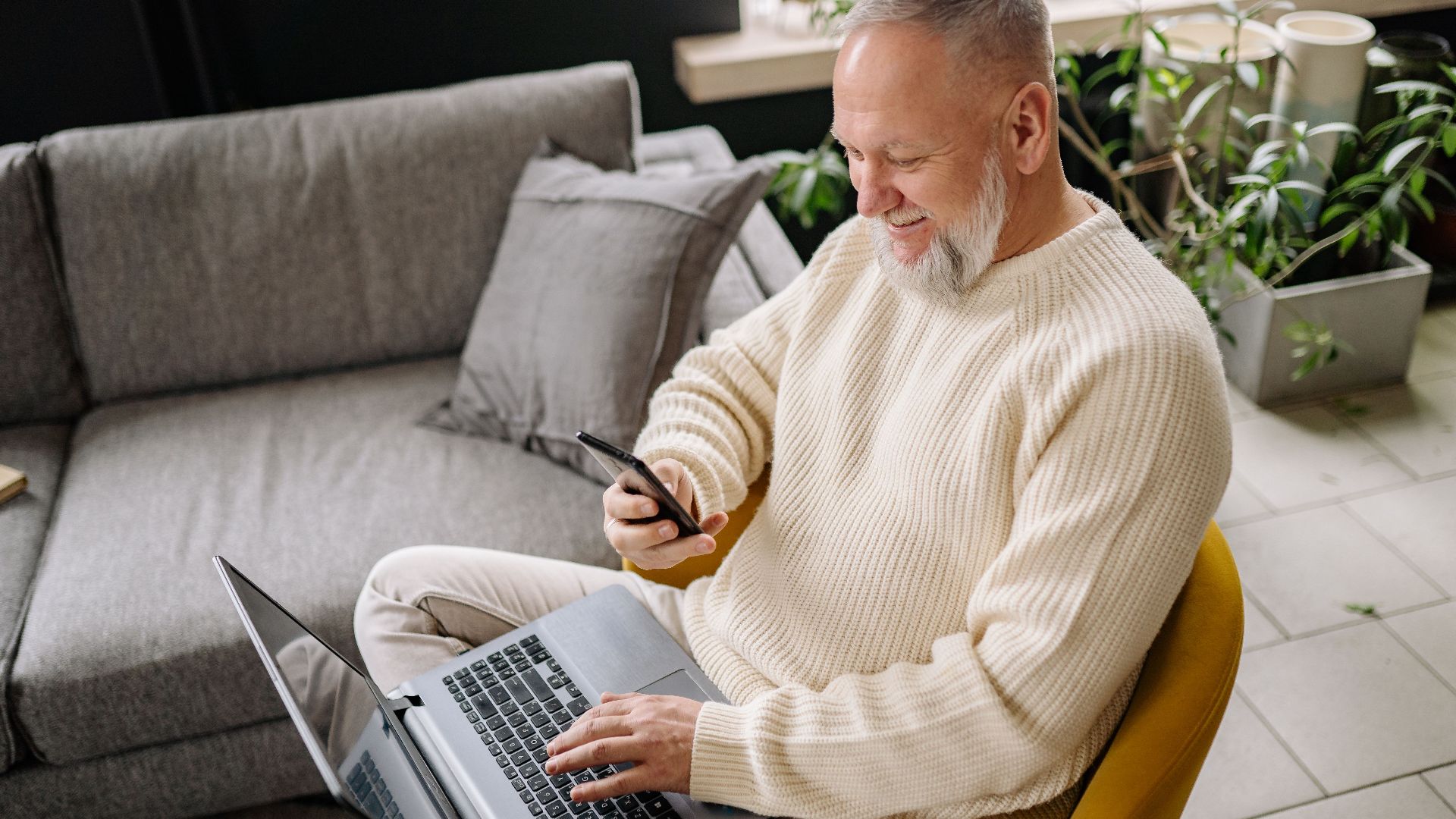 A smiling elderly man sits comfortably indoors, using both a phone and laptop, with a cozy ambiance.