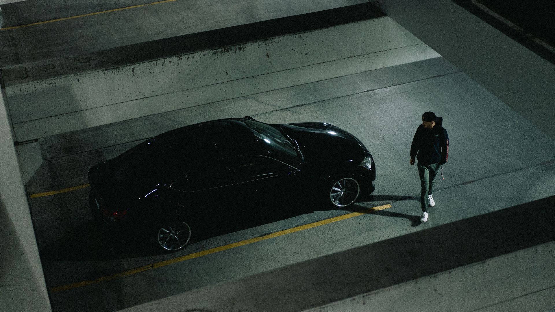 High angle view of a man walking beside a parked black car in a dimly lit parking garage.
