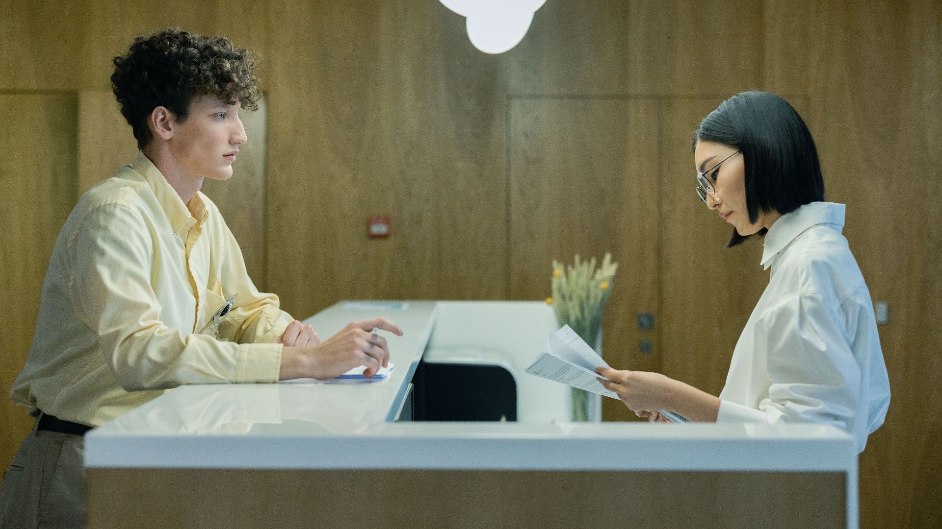 A young man and woman interacting at a modern office reception desk indoors.