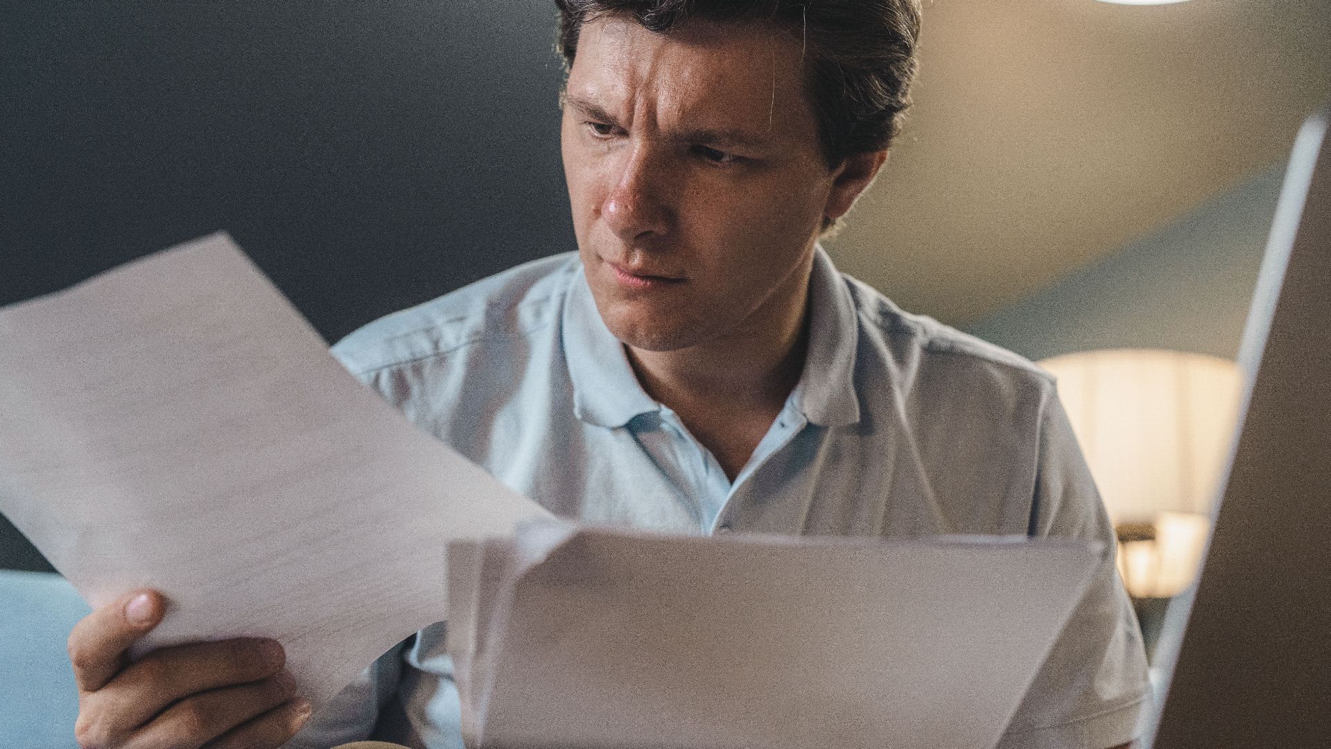 A man intently examines papers, seated indoors under warm lighting, focusing on his work.