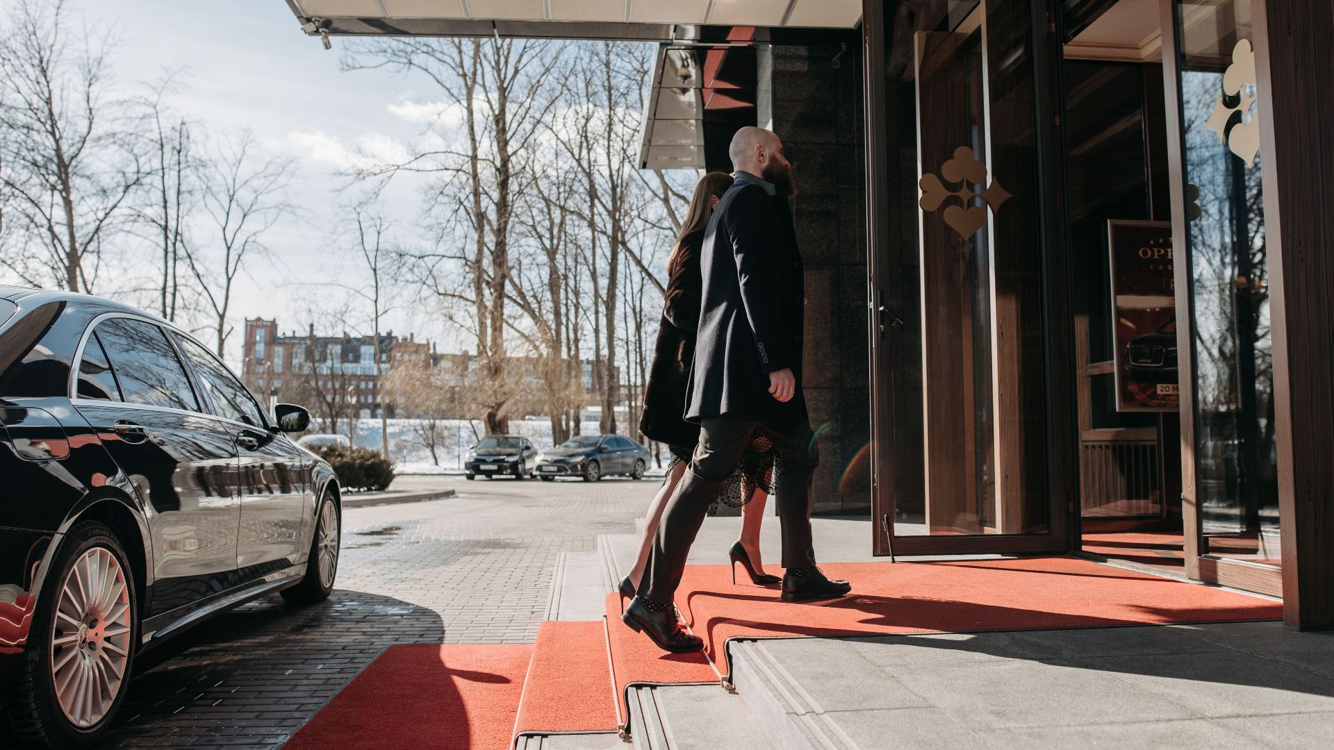 A man and woman elegantly dressed enter a luxury hotel, walking on a red carpet.