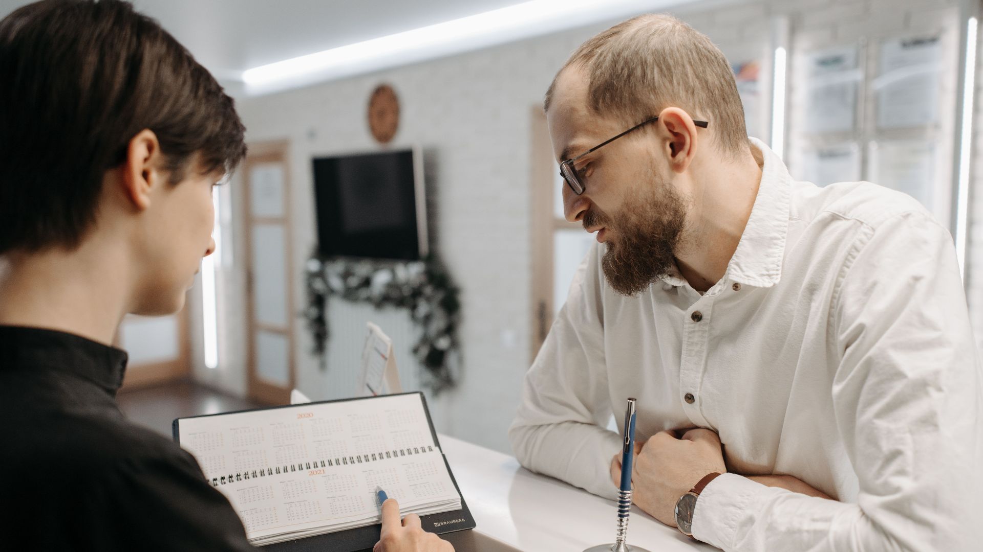 A receptionist and client converse over an appointment book at a clinic reception desk.