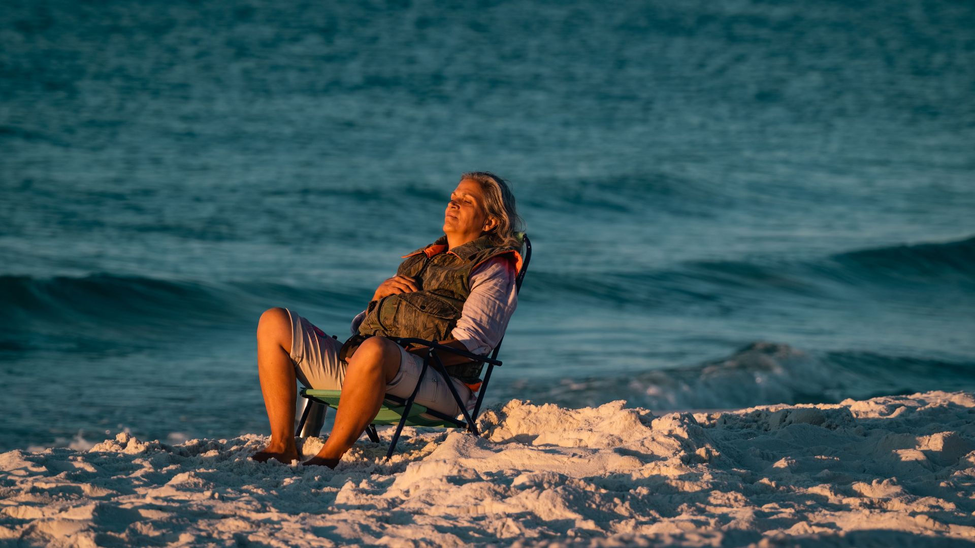 A woman enjoys a serene moment on a beach chair by the ocean at sunset.
