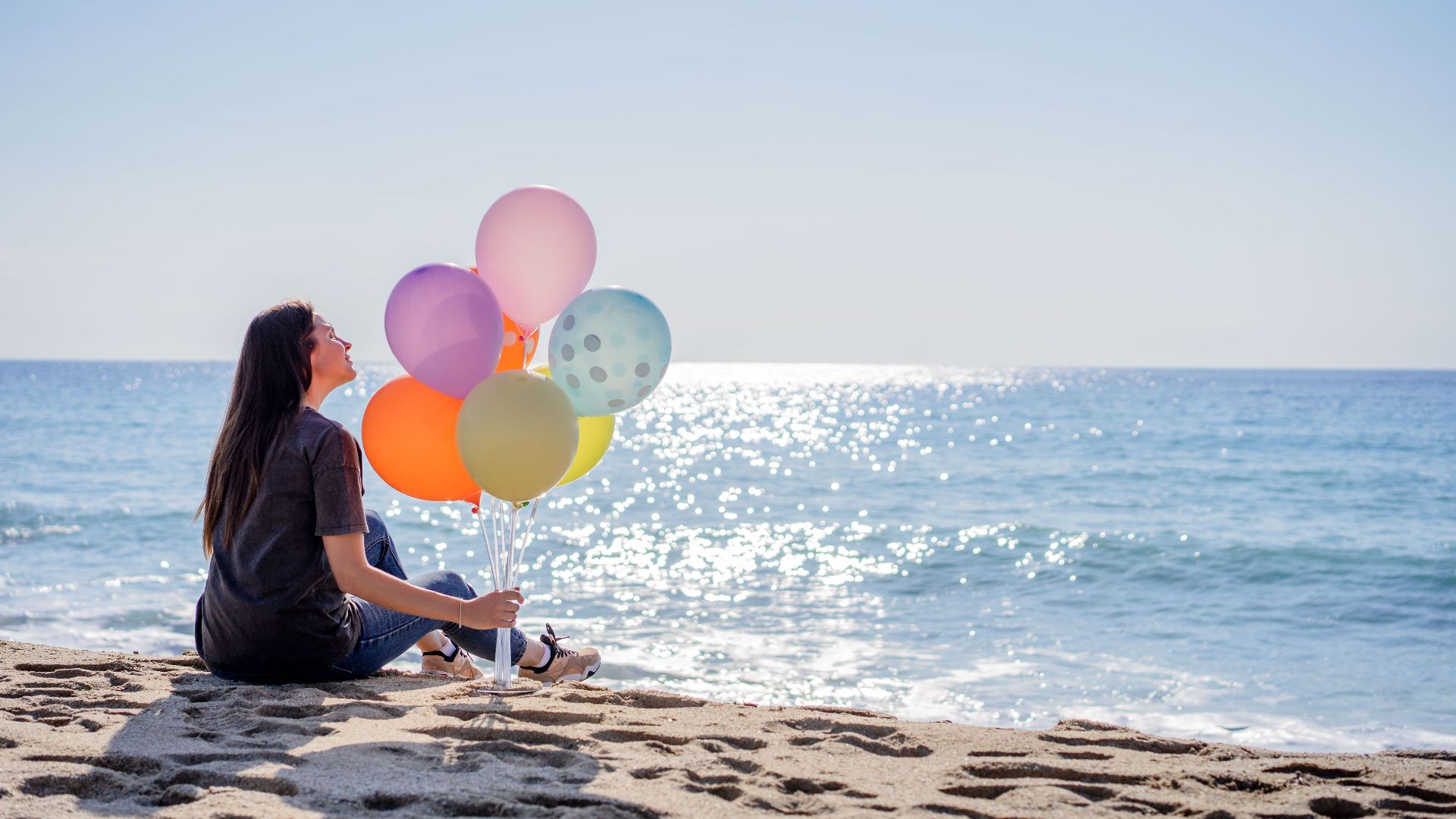 A woman enjoying a peaceful day at the beach holding colorful balloons by the sea.