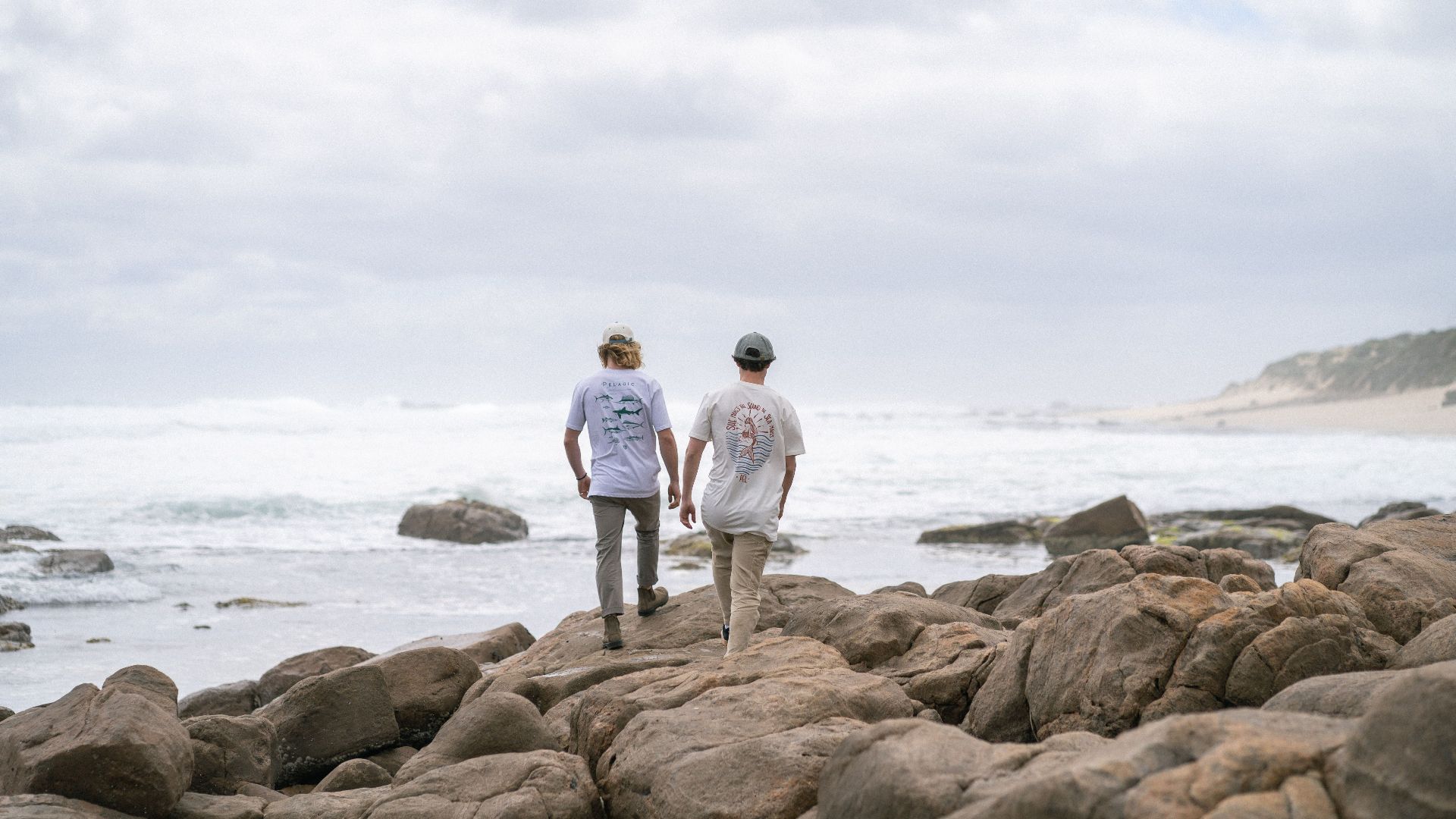 Two men walk on rocky beach with waves and clouds in the horizon, creating a serene outdoor scene.