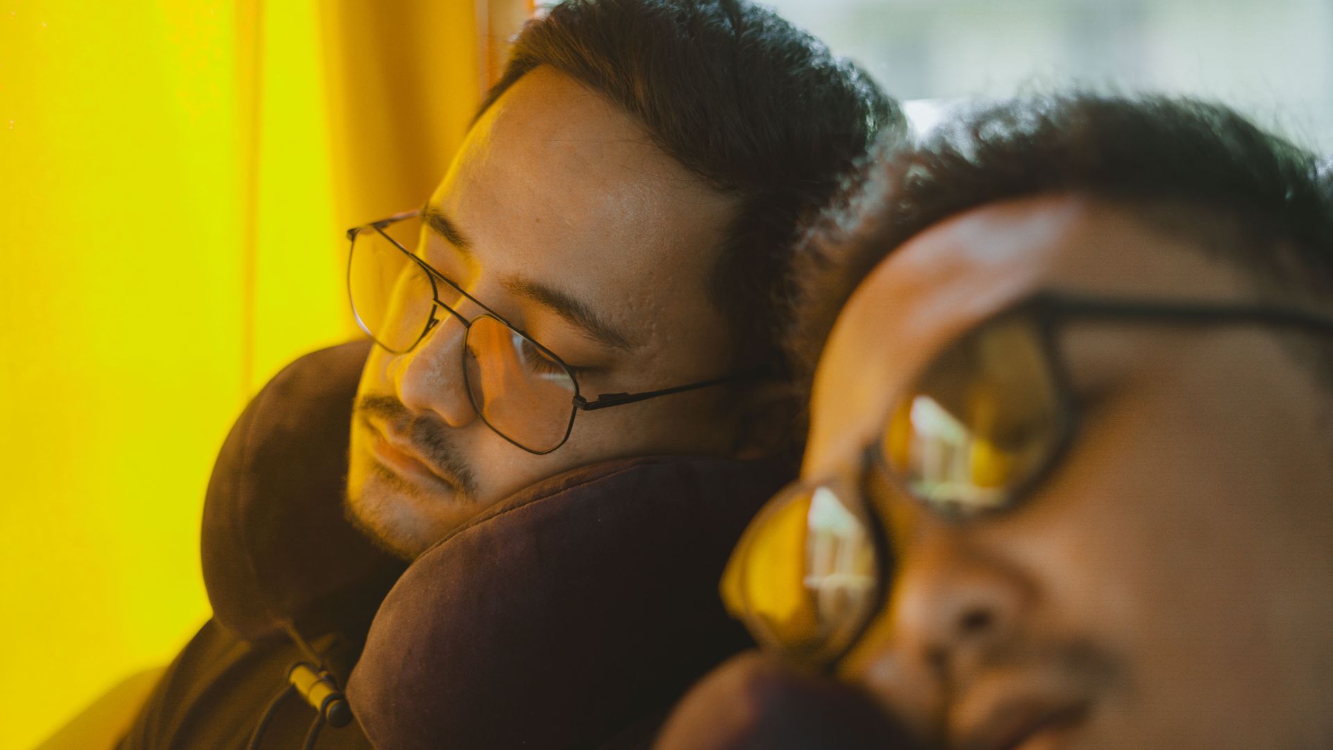 Two men wearing eyeglasses sleep on a bus, using neck pillows for comfort during the journey.