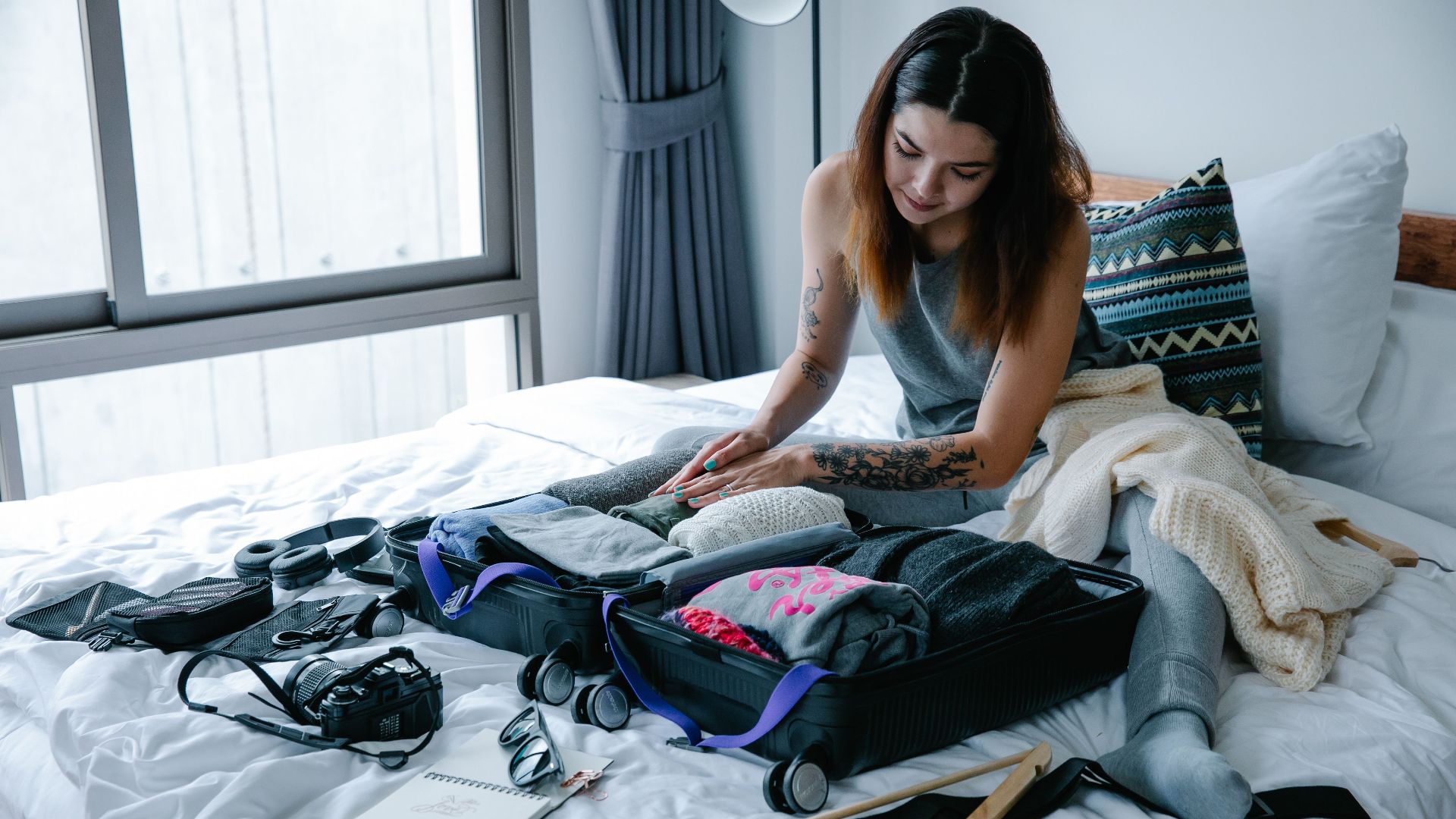 Woman sitting on bed packing suitcase with clothes and travel essentials.