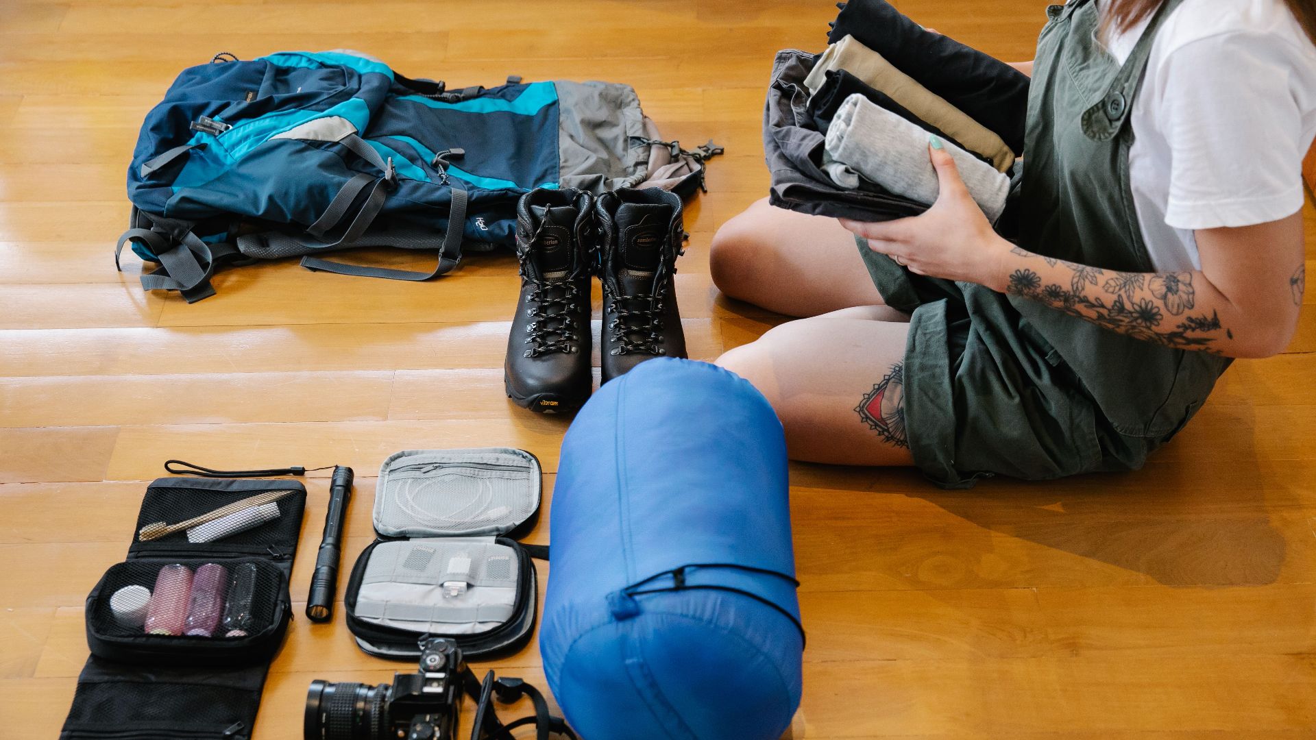 Woman organizing camping gear and clothes on wooden floor indoors, preparing for an adventure.