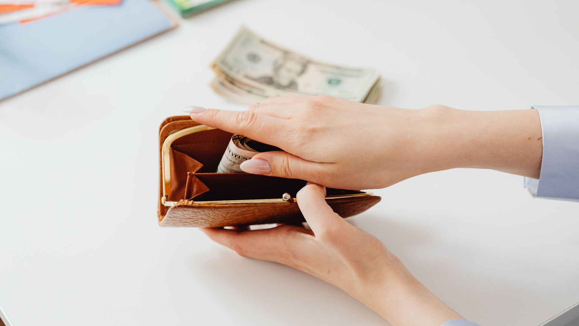 Close-up of hands placing cash into a leather wallet with bills on table.