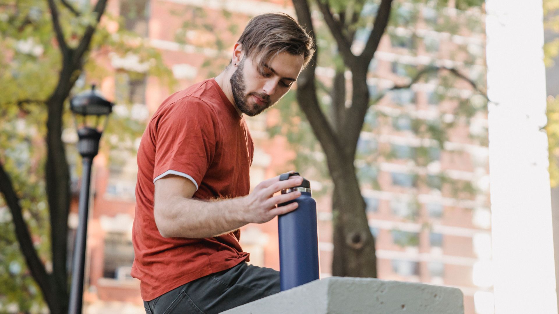 Low angle side view of young unshaven male athlete with bottle resting on fence in town