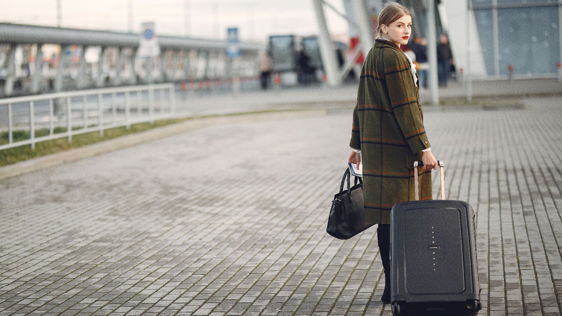 Full length of stylish female traveler in warm coat walking with suitcase and bag looking at camera after arrival against airport terminal