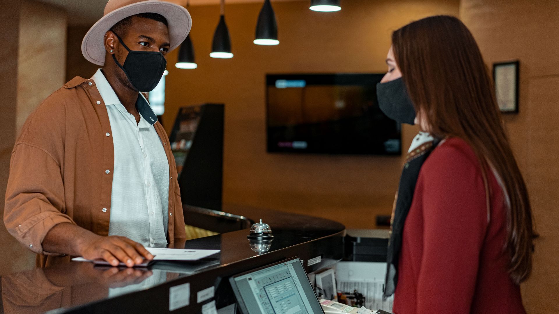 A man and woman in face masks checking in at a hotel reception, emphasizing safety protocols.