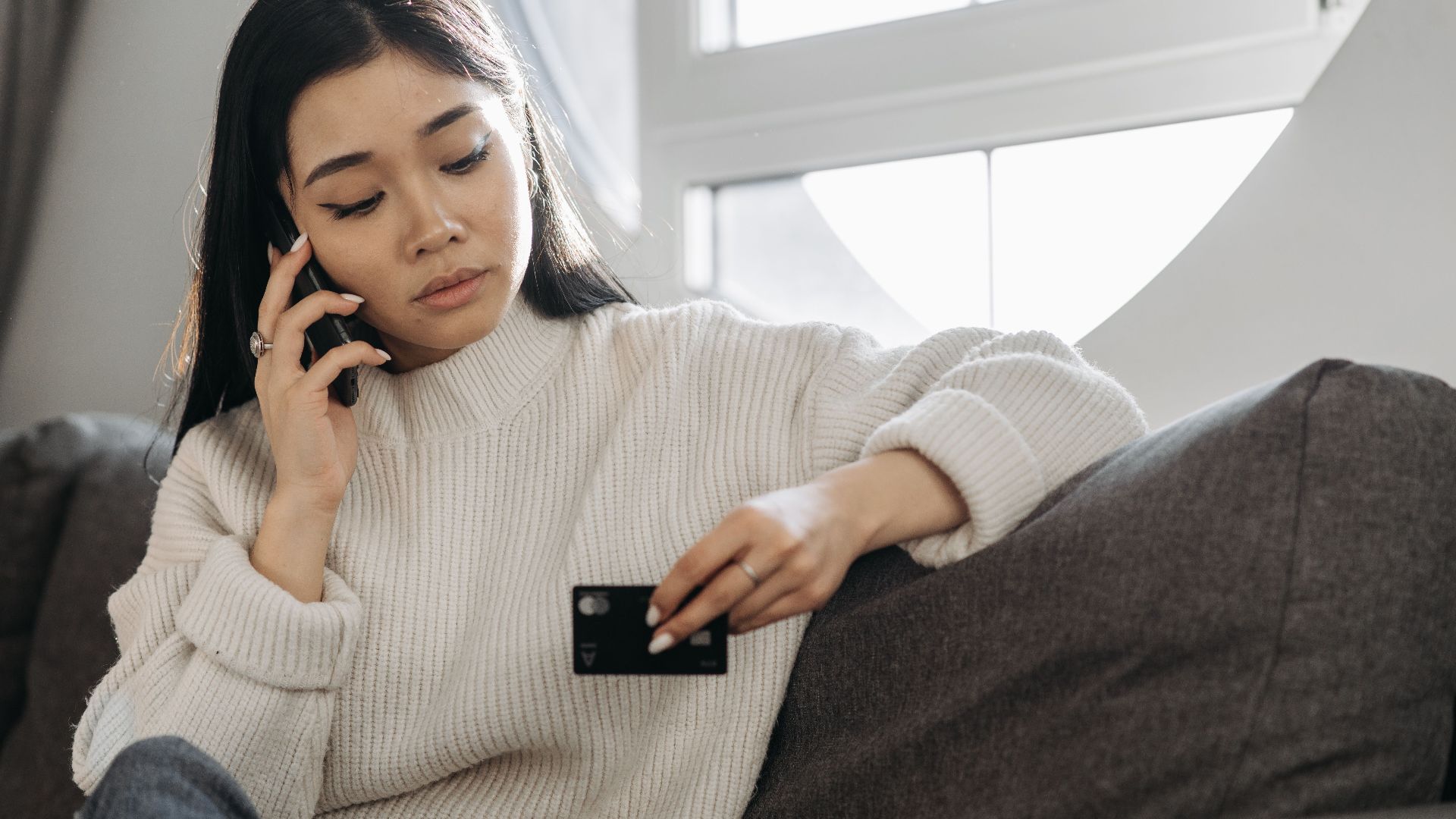 A woman sits on a sofa, multitasking with a phone and credit card, focusing on online shopping.