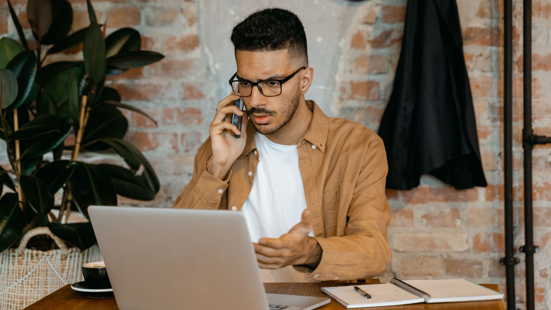A man in eyeglasses works on a laptop while talking on the phone in a stylish brick-walled office.