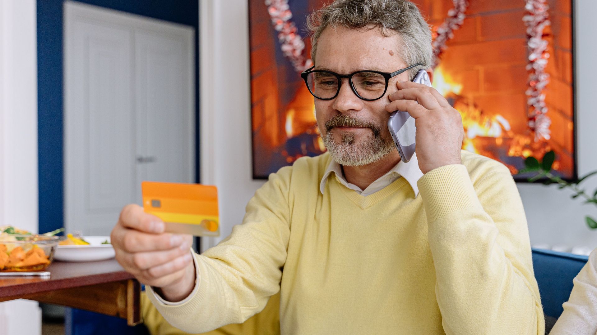 Elderly man using phone while holding a credit card indoors near a cozy fireplace.
