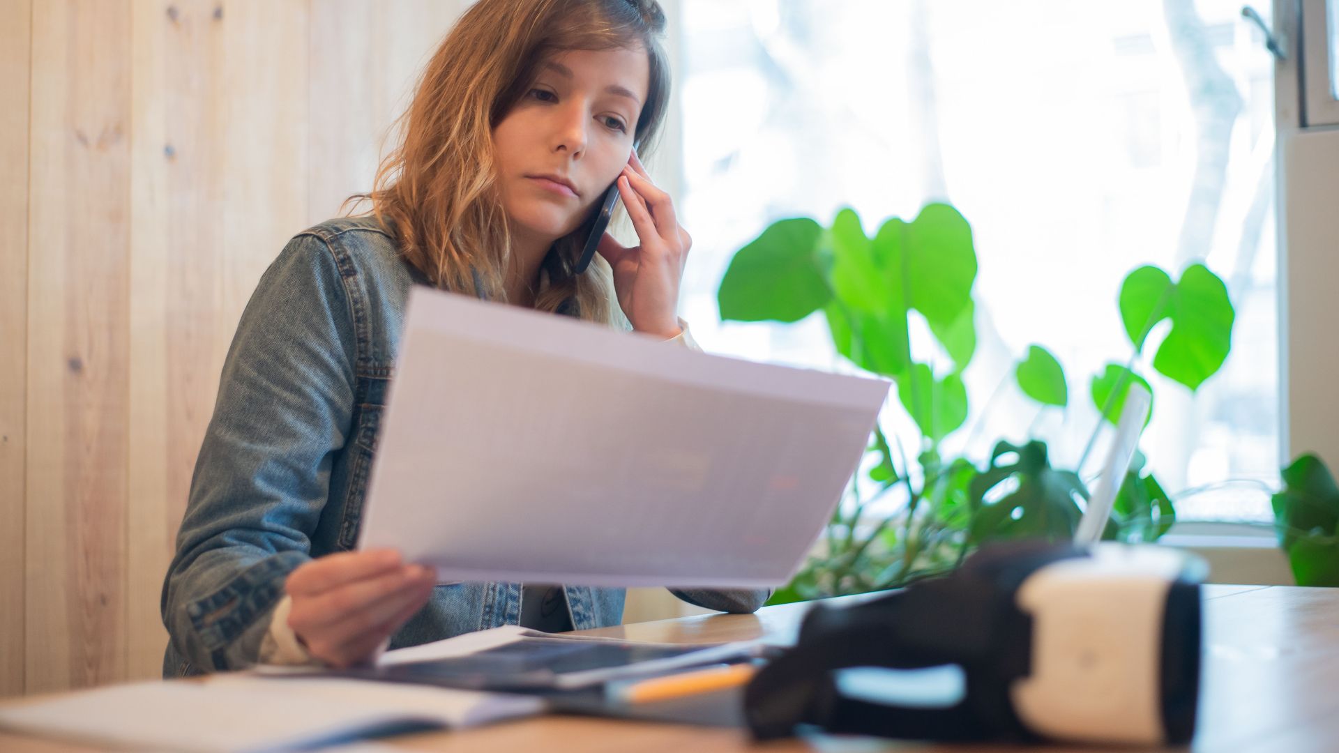 Businesswoman reviewing paperwork while on a call at a modern office workspace.