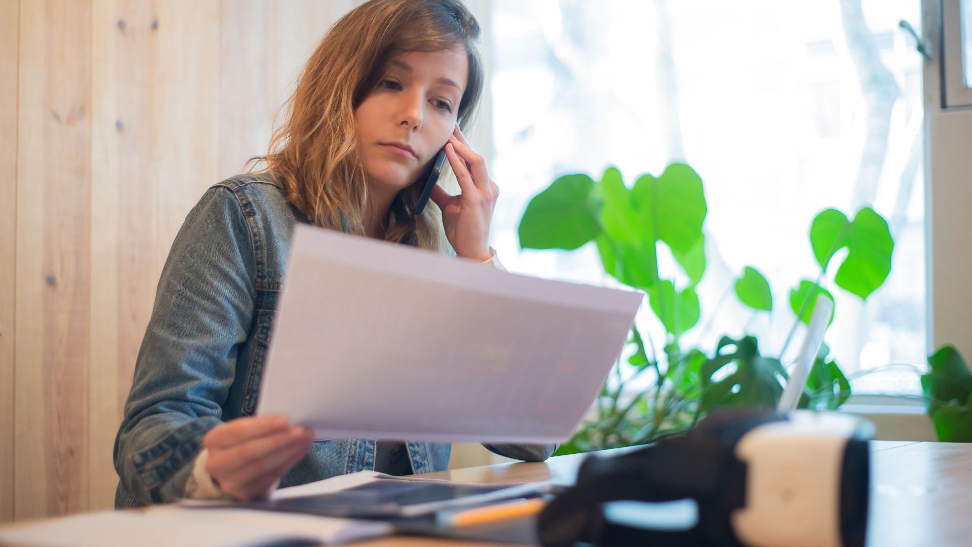 Businesswoman reviewing paperwork while on a call at a modern office workspace.