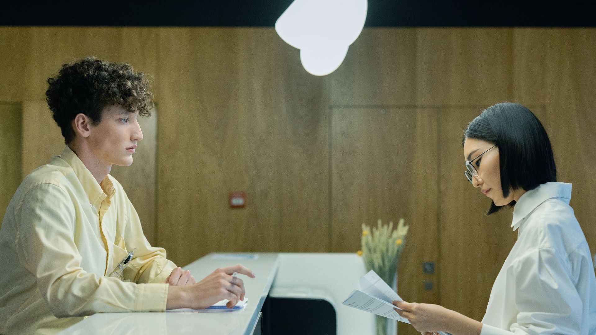 A young man and woman interacting at a modern office reception desk indoors.