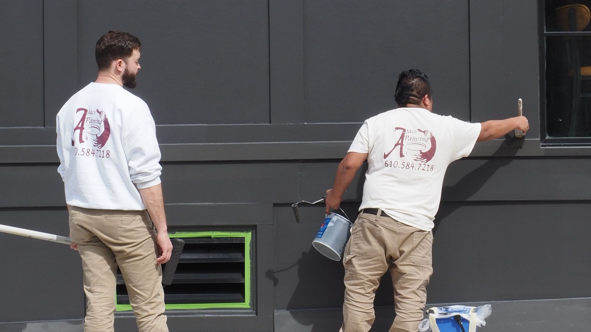 Two men painting a large dark wall in an urban setting, wearing work uniforms.