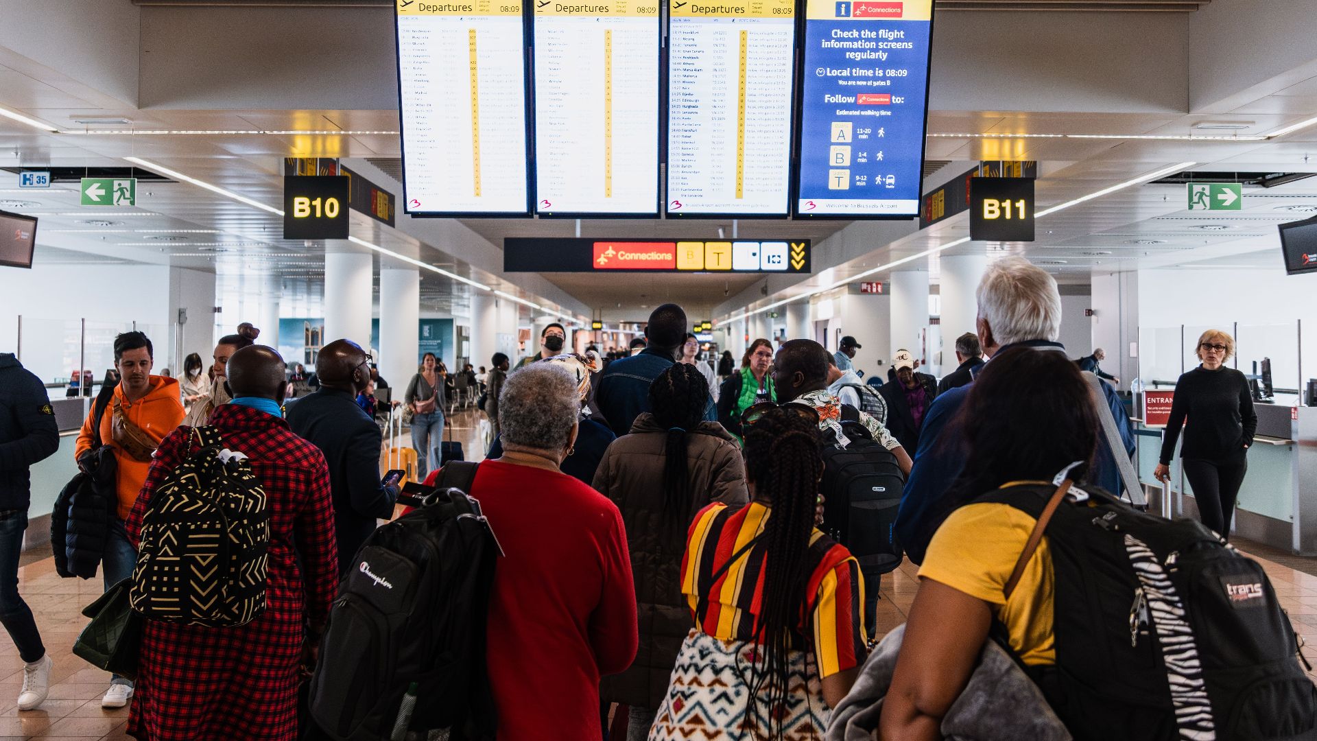 Travelers lining up in a busy airport terminal with baggage under flight information screens.