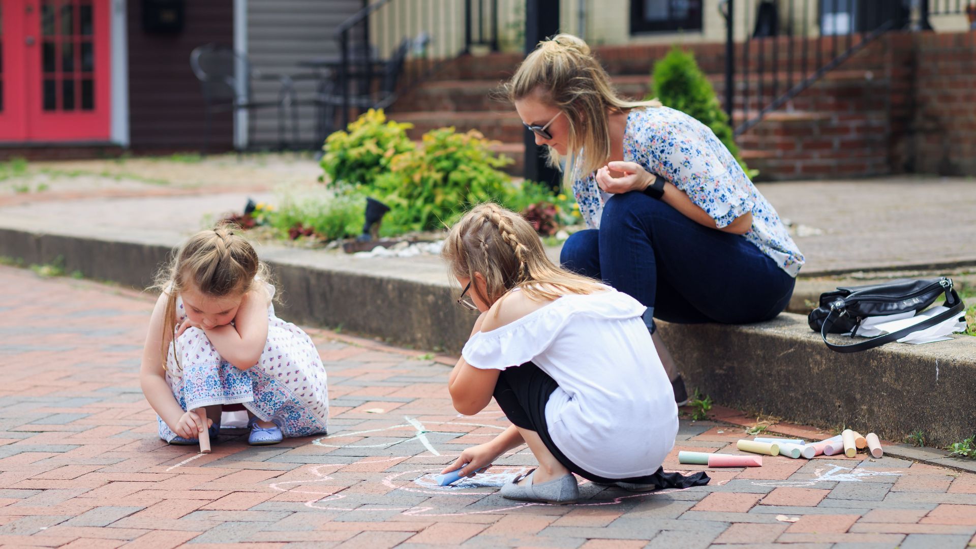 Two children drawing with chalk on a brick sidewalk, watched by a woman. Outdoor fun and creativity.