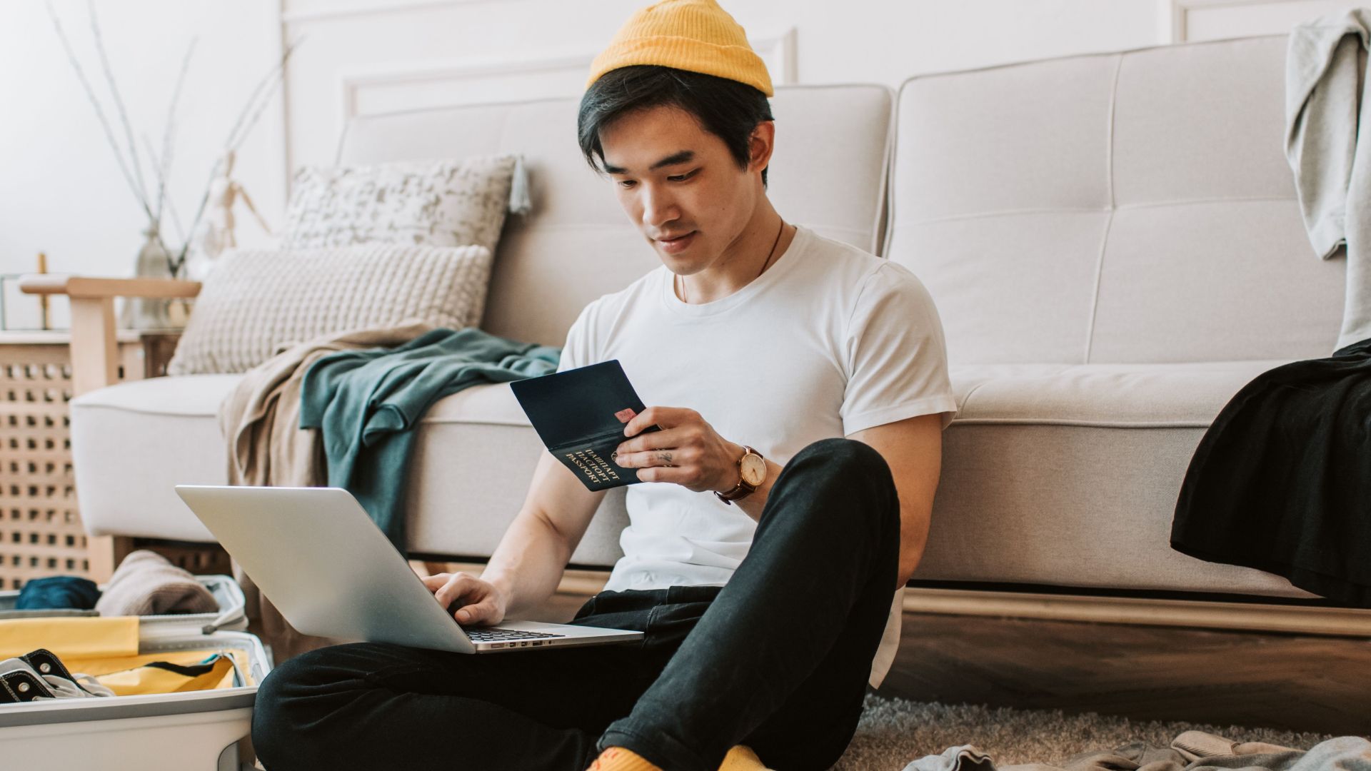 A young man in a messy living room preparing for travel with a laptop and passport.