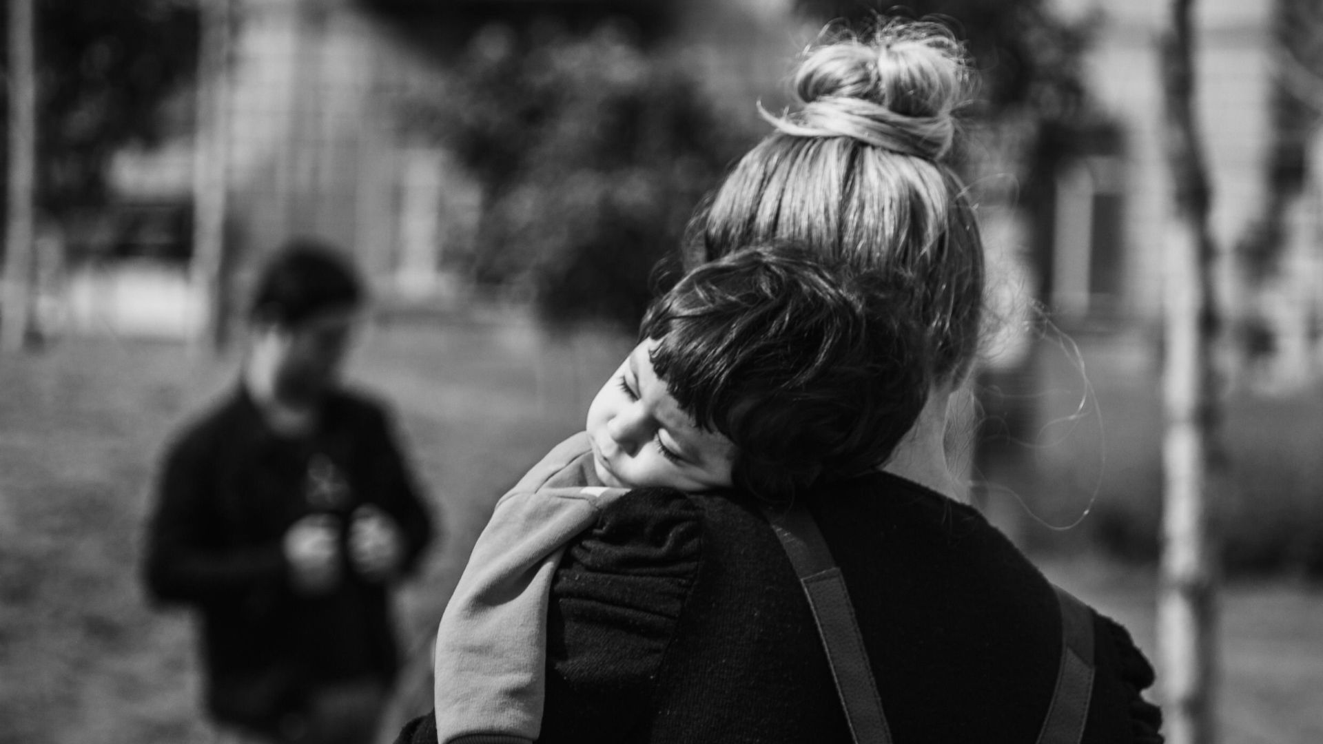 A serene black and white photo of a mother carrying her sleeping child outdoors.