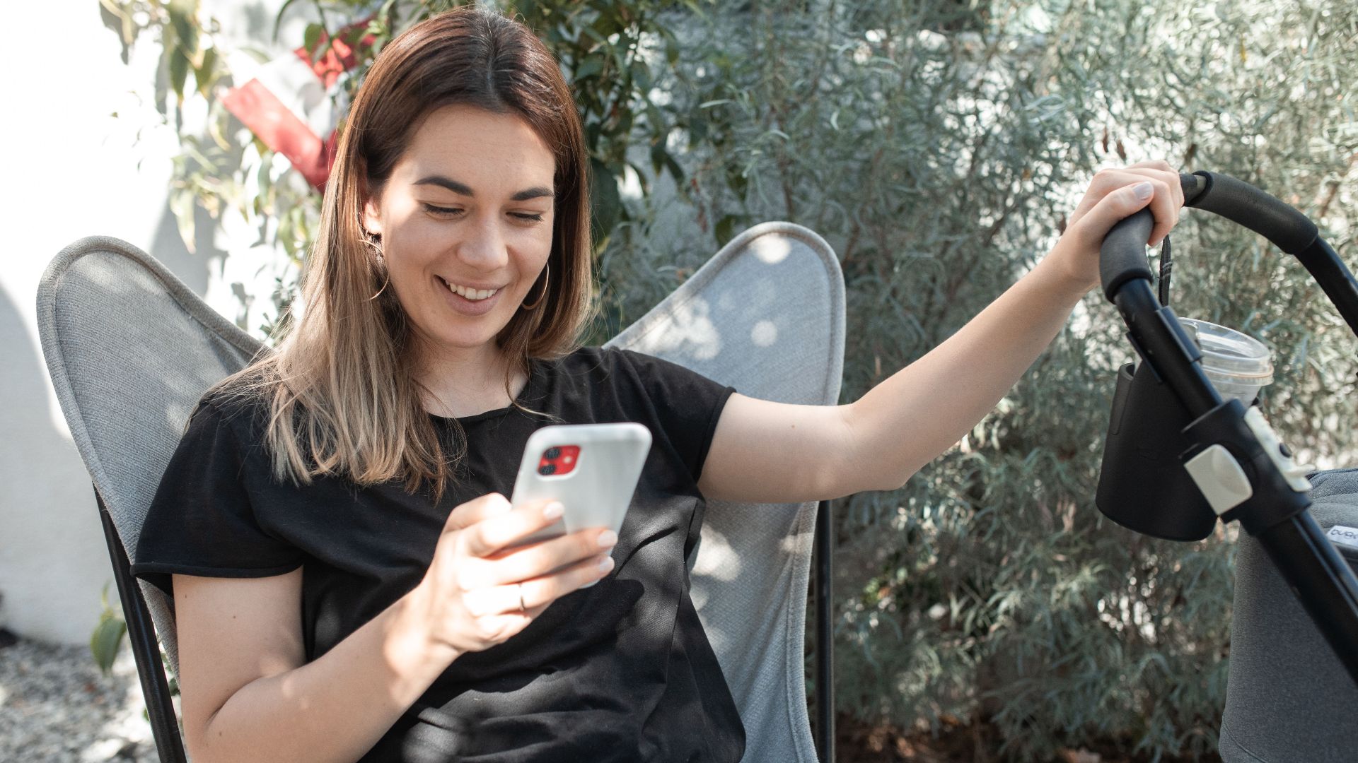 Woman seated outdoors, smiling while using smartphone beside a stroller.