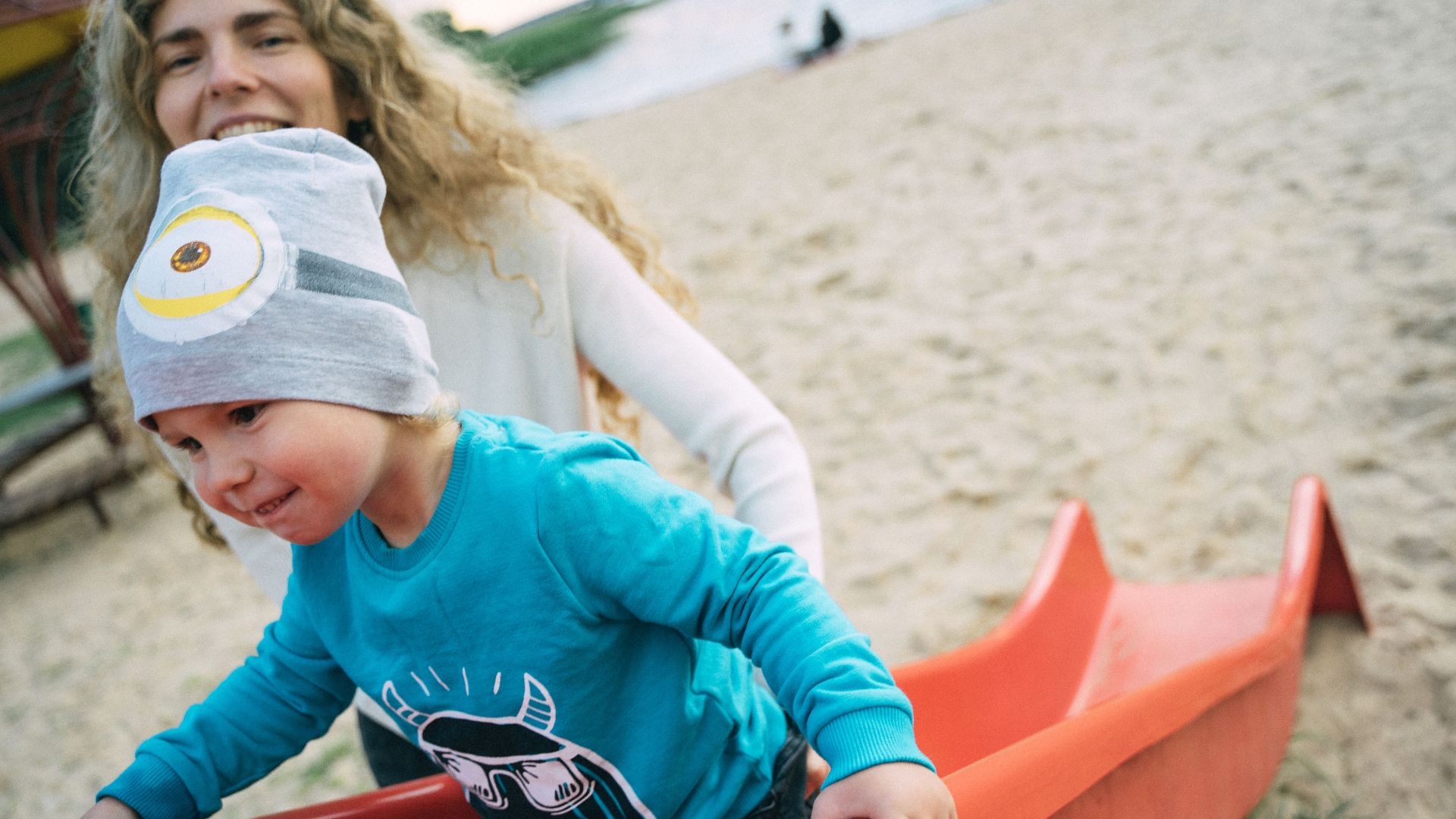 A mother guiding her child down a slide at the beach playground. Fun family bonding moment.