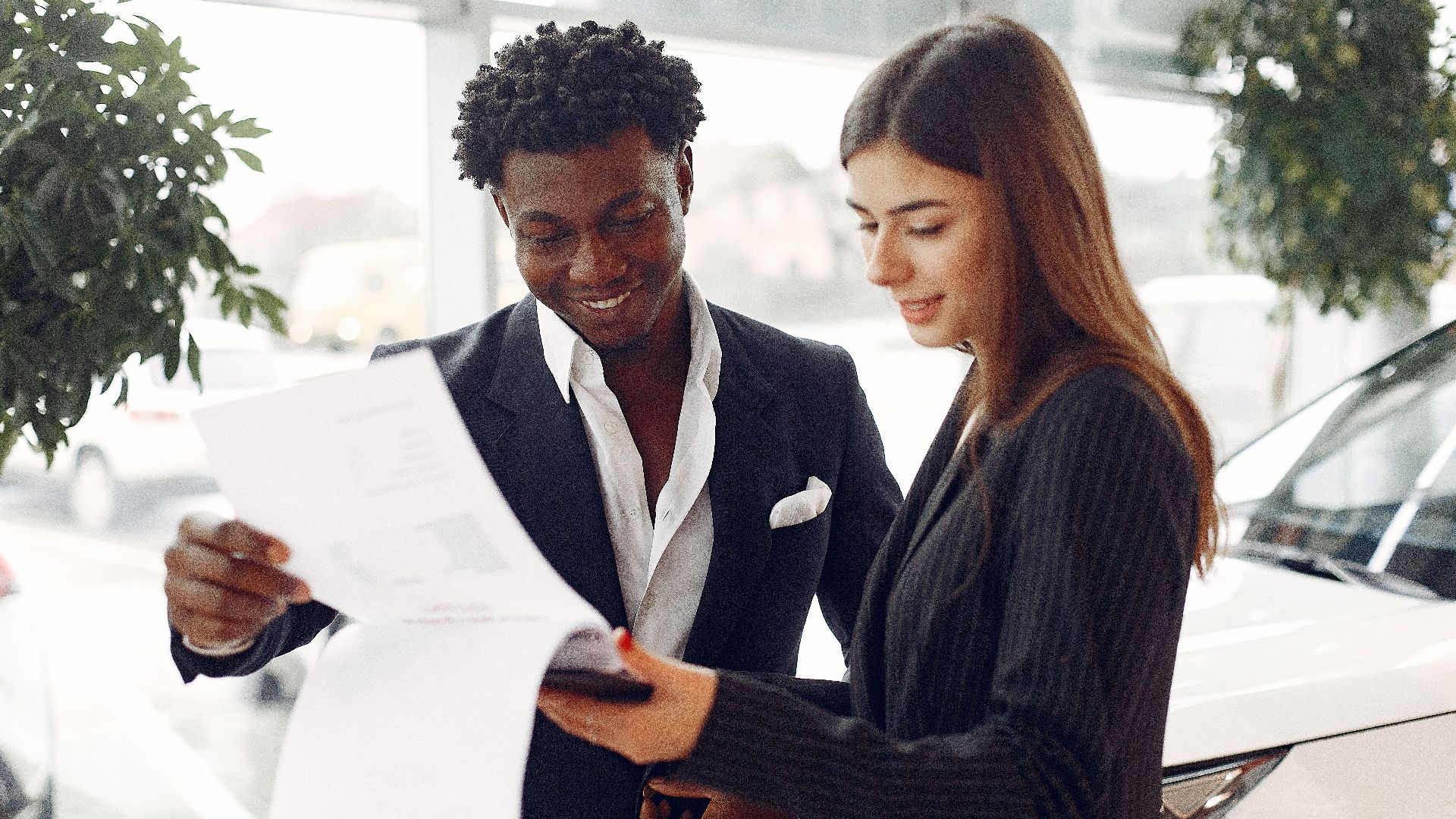 Cheerful smiling multiethnic businessman in classy suit and professional elegant female consultant standing close and reading contract details in car showroom