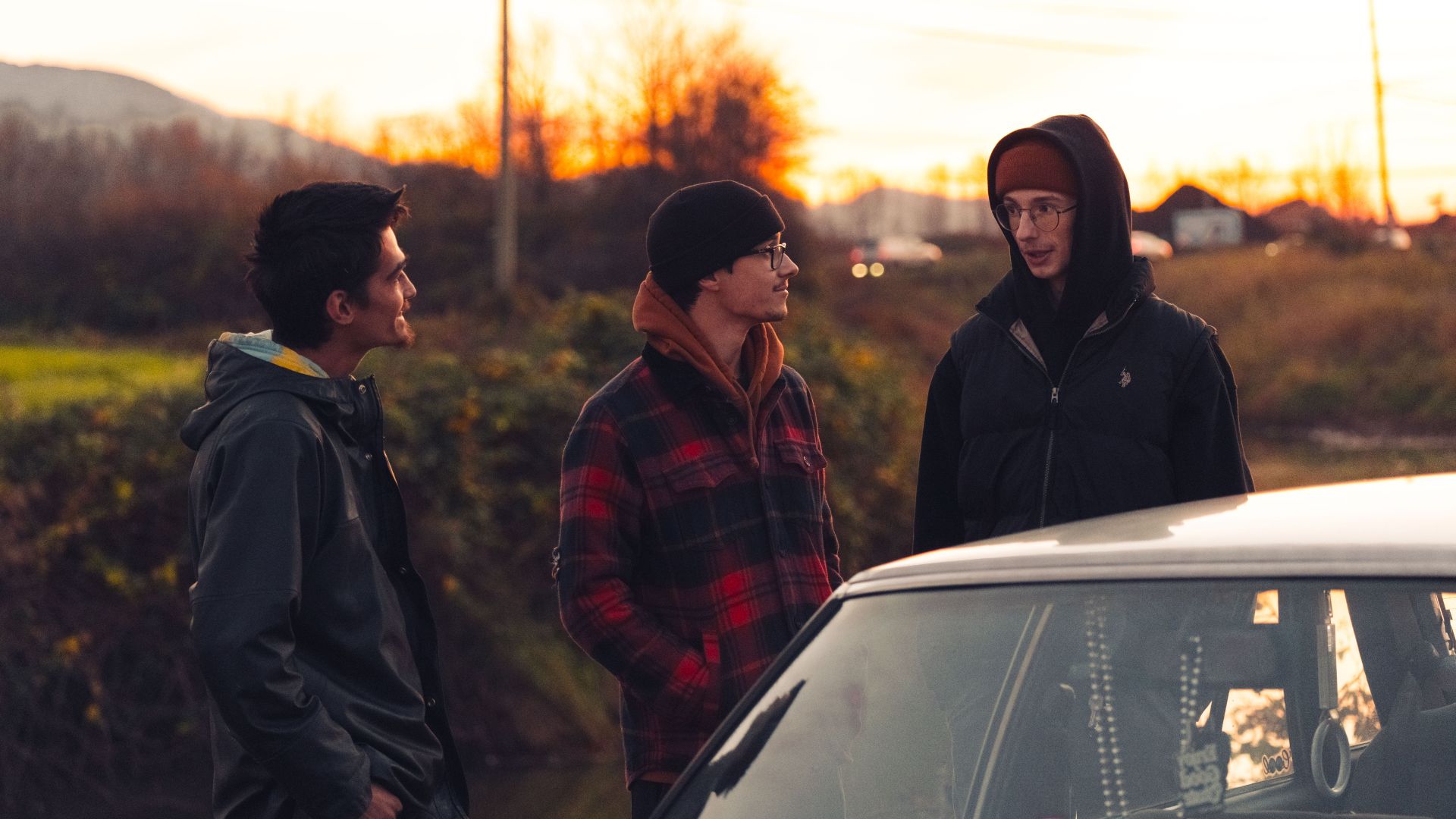 Three young men enjoy a sunset chat outdoors, bundled in warm attire beside a car.