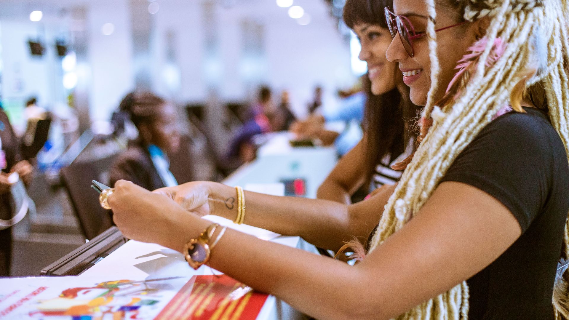 Two stylish women with unique hairstyles smiling at an indoor counter in a lively setting.