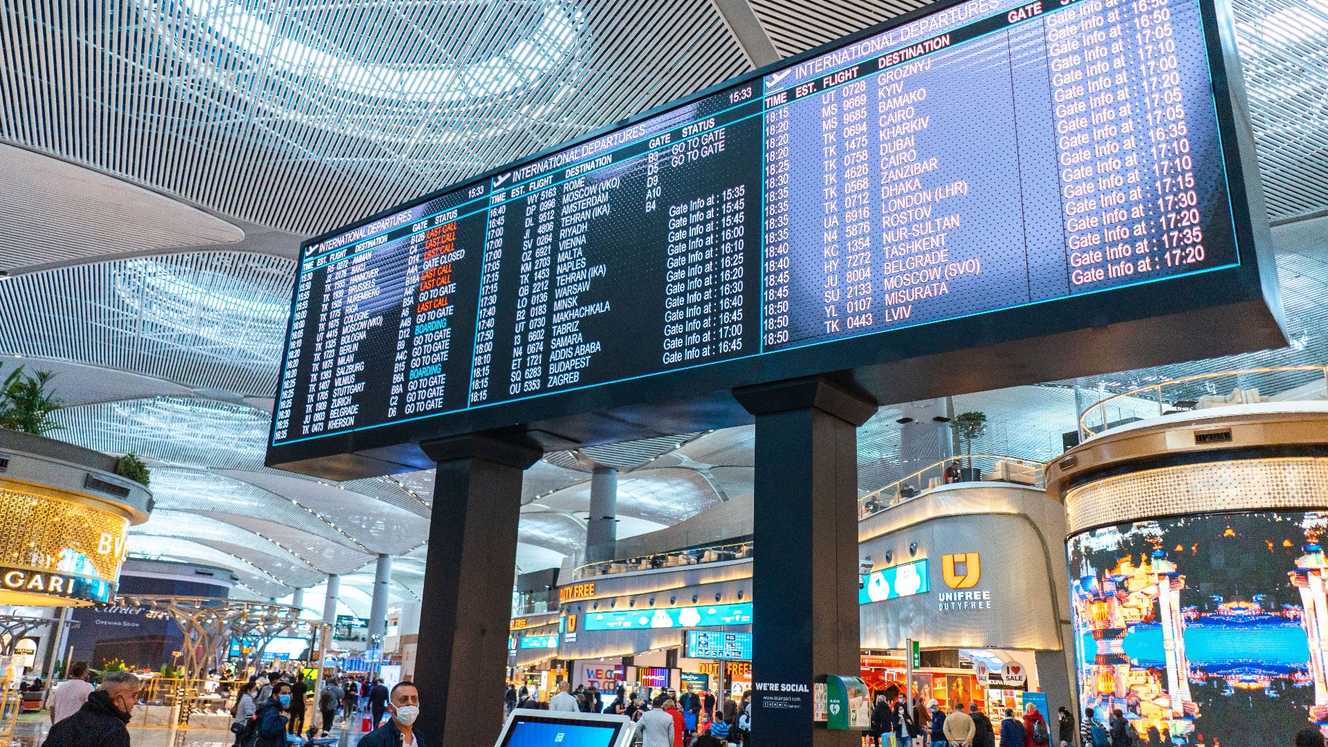 Passengers walk through Istanbul Airport's departure area, surrounded by stores and flight information displays.