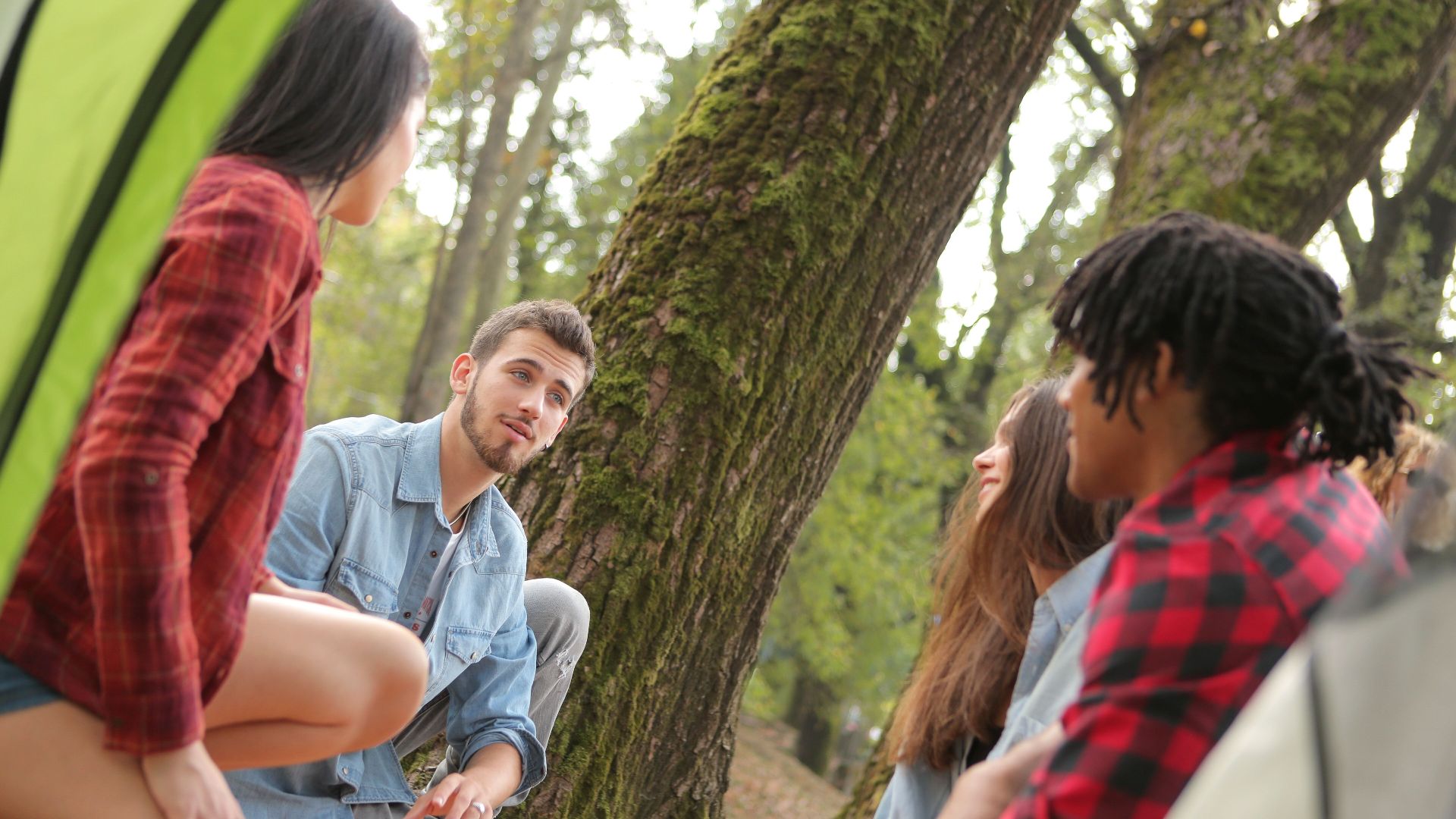 Group of young people in casual clothes sitting on plaid around near trees with moss while talking and looking at each other
