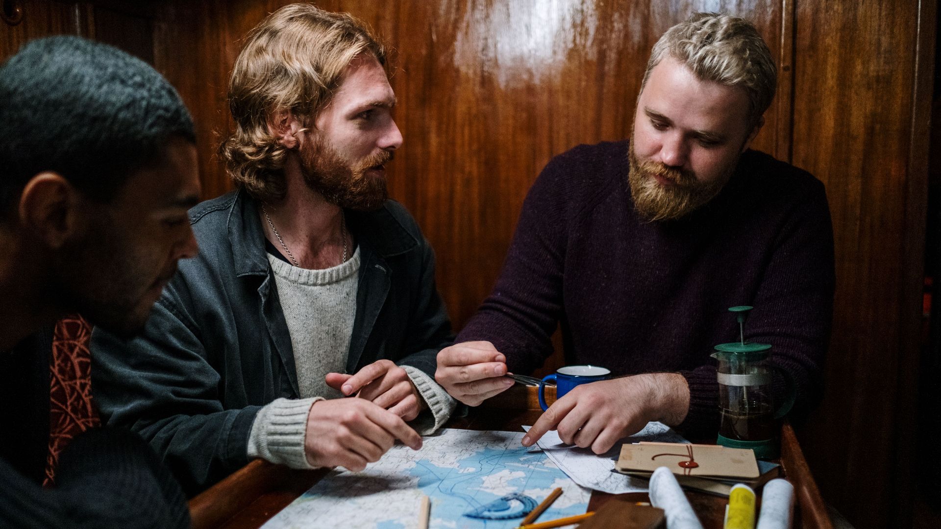Three men planning a nautical journey using maps indoors, discussing routes and navigation.