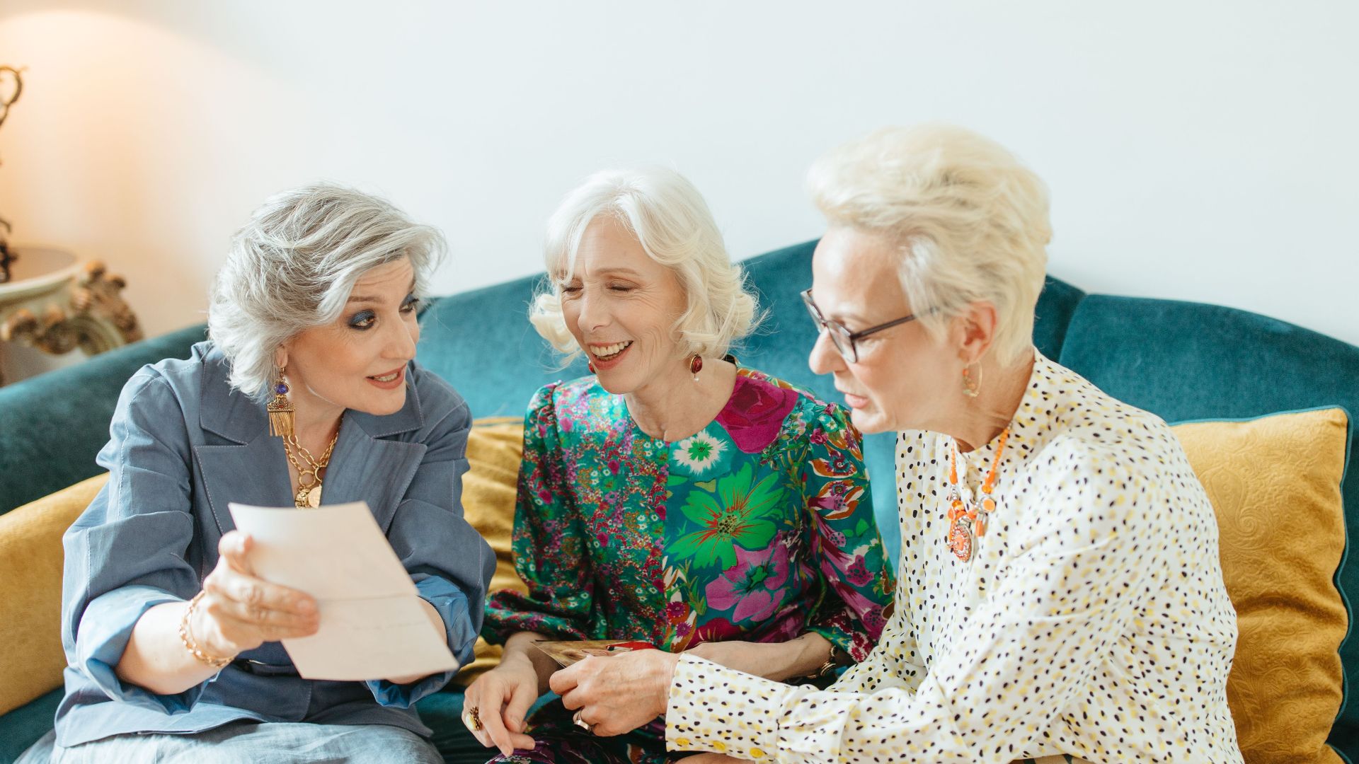 Three senior women joyfully reminiscing over old photos on a cozy couch.