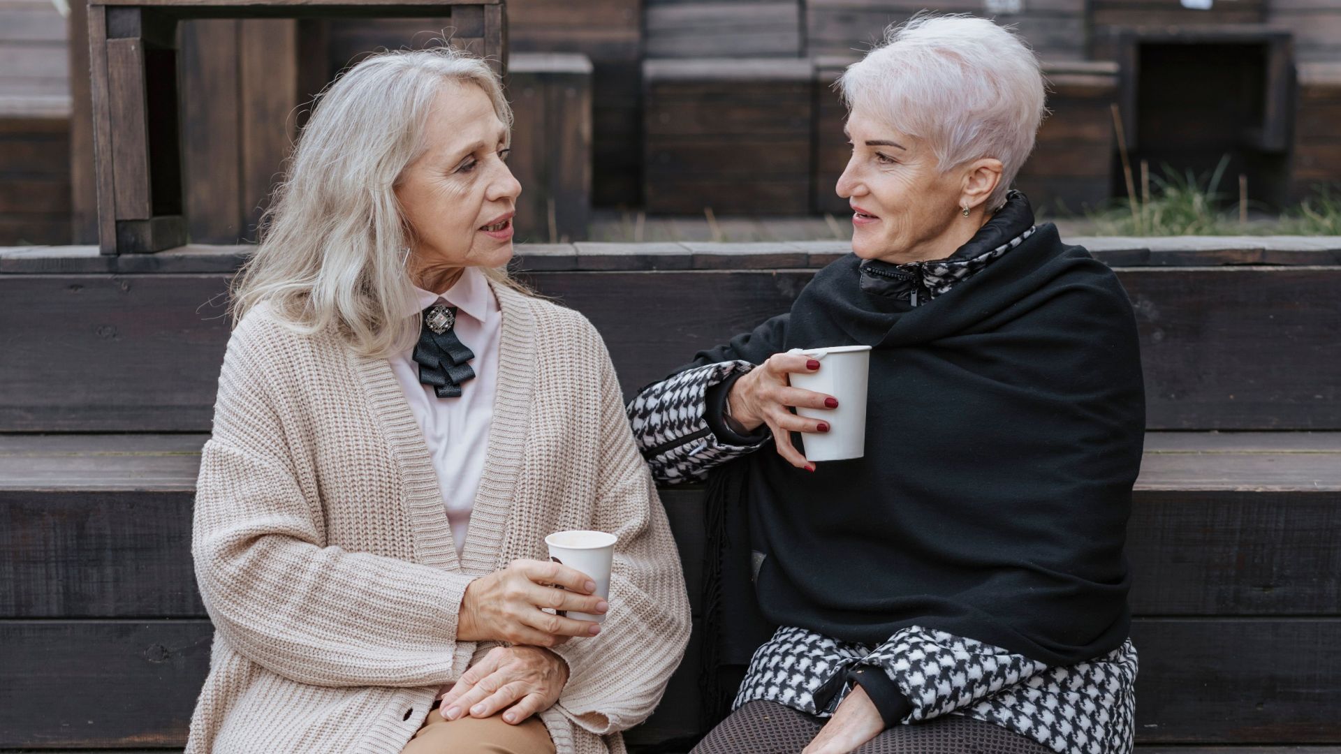 Two senior women sitting on a bench, sharing coffee and conversation in a cozy outdoor setting.