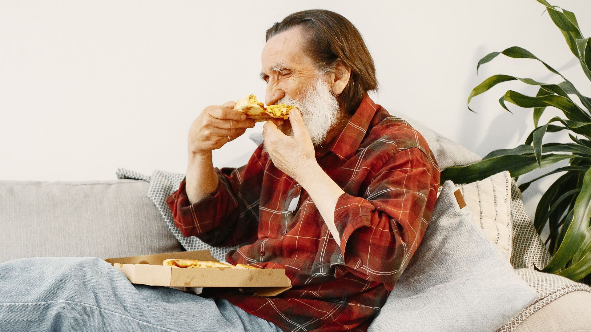 Senior man savoring a slice of pizza while lounging on a comfortable couch at home.
