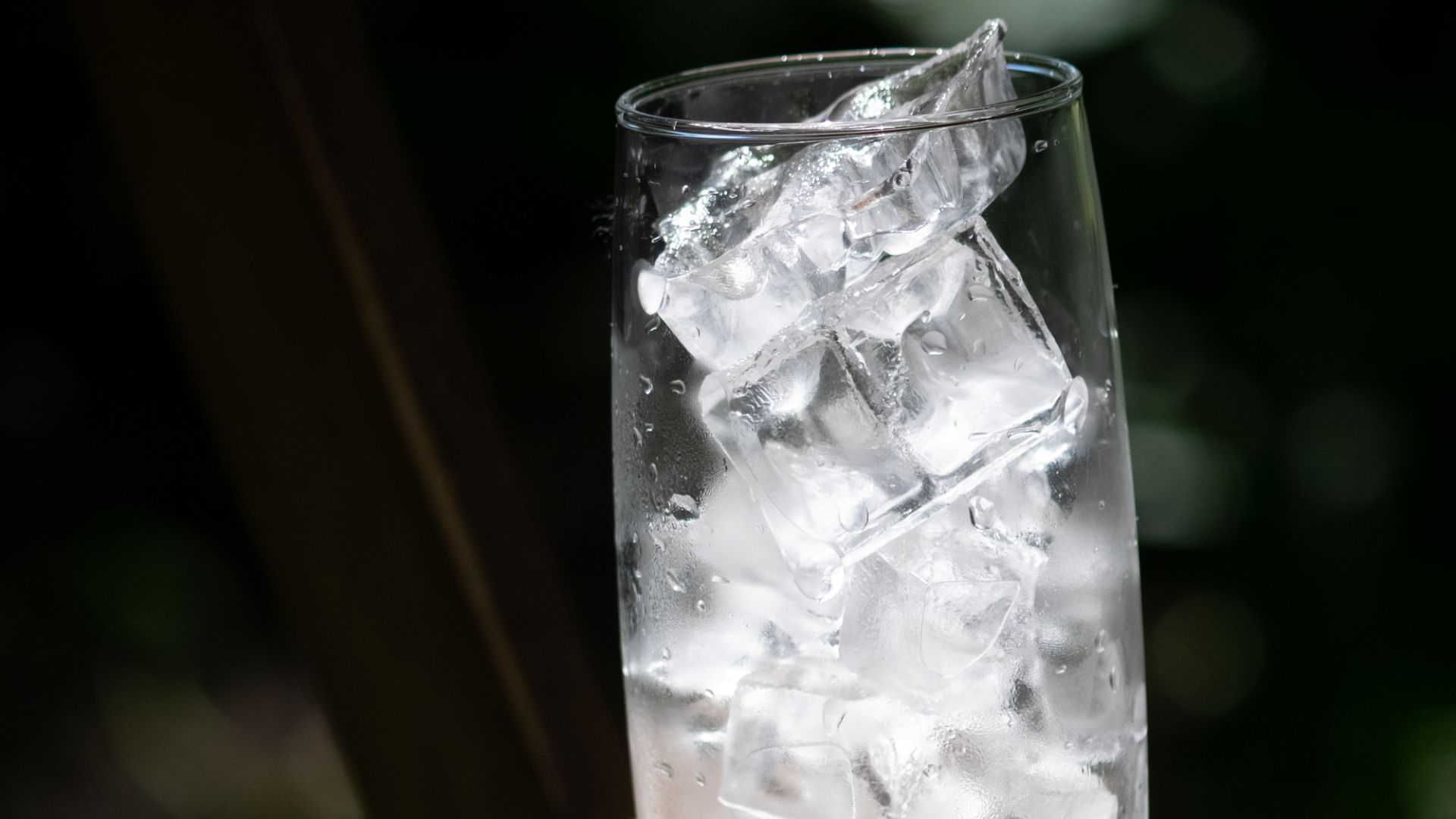 A tall glass filled with ice cubes resting on a wooden table outdoors in sunlight.