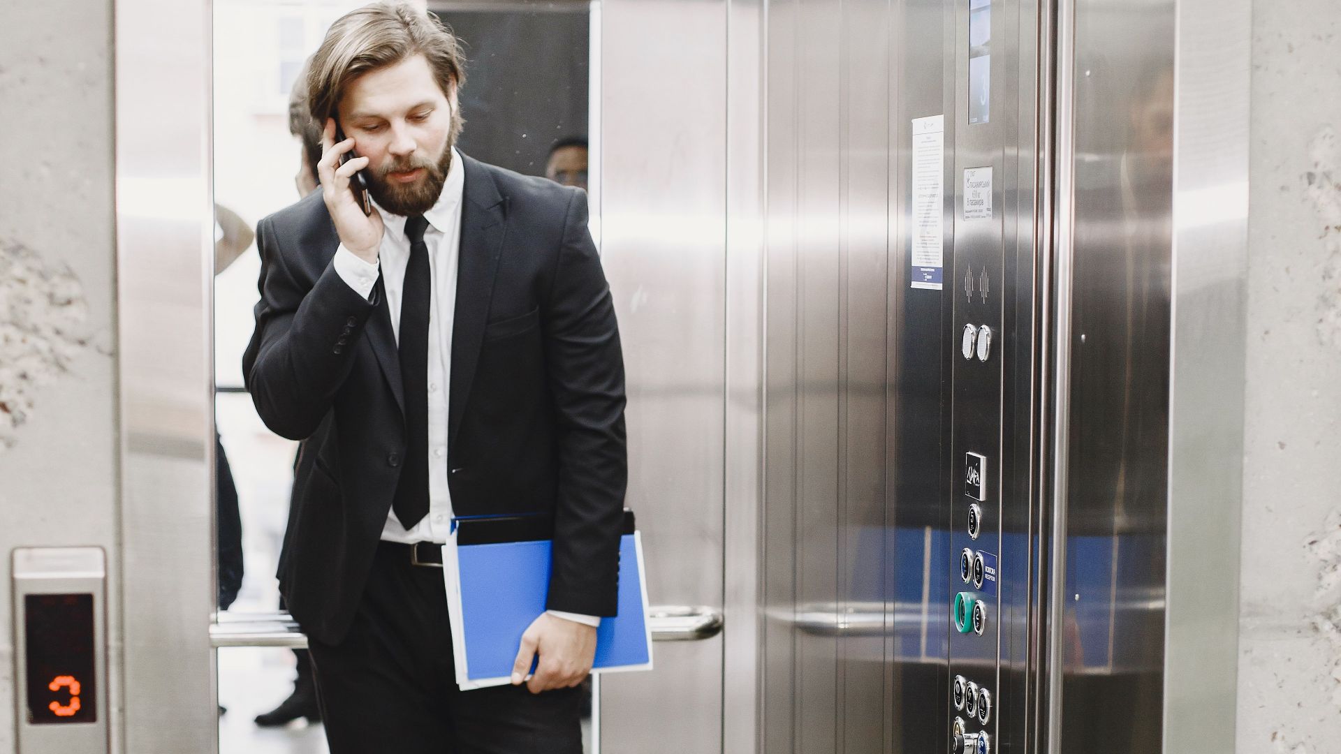 A businessman in a suit talking on his phone inside an elevator holding documents.