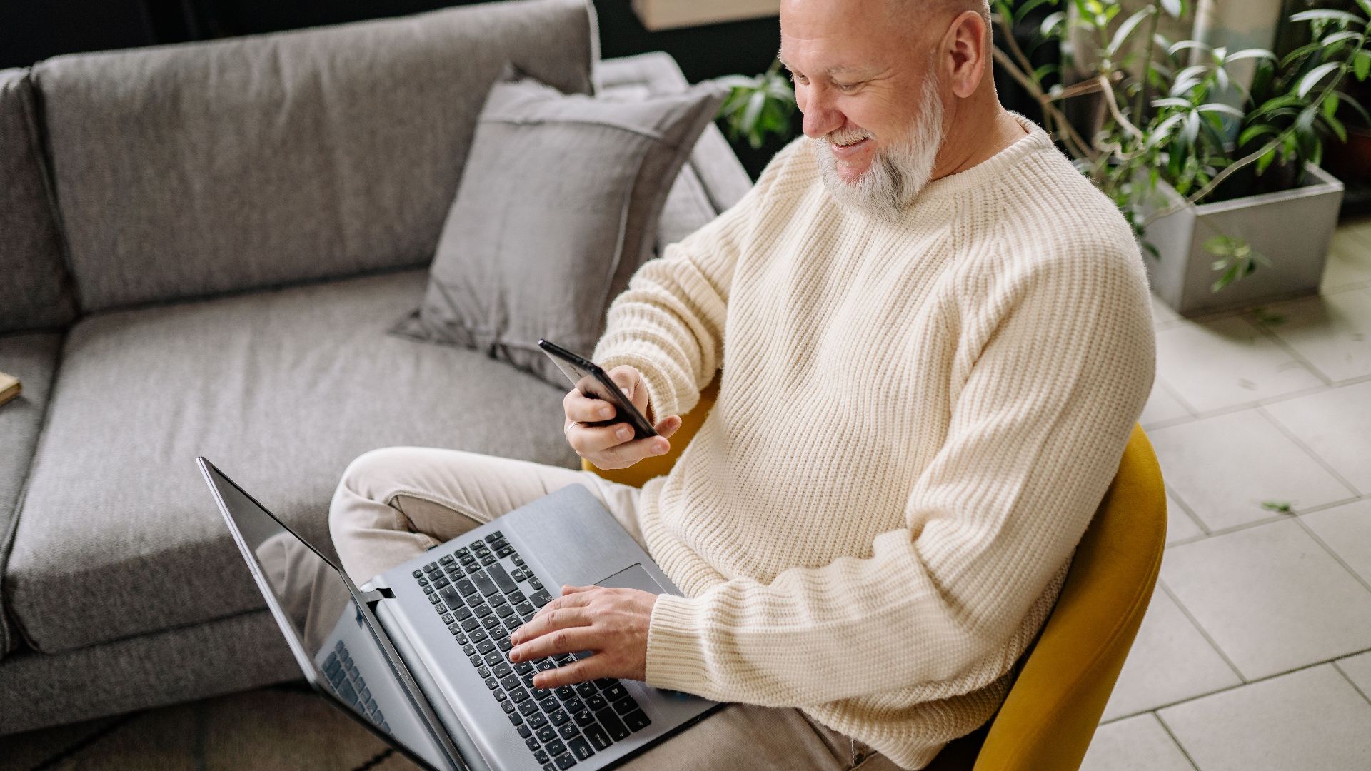A smiling elderly man sits comfortably indoors, using both a phone and laptop, with a cozy ambiance.