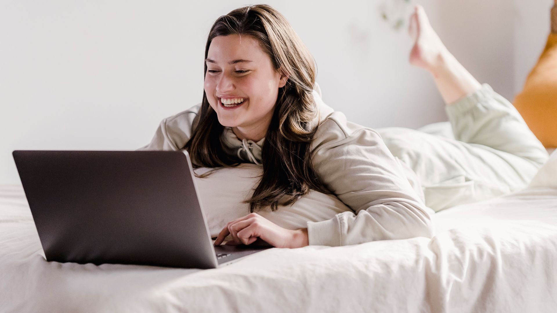 Happy young female smiling and browsing internet on modern netbook while resting on comfortable bed in light bedroom at home