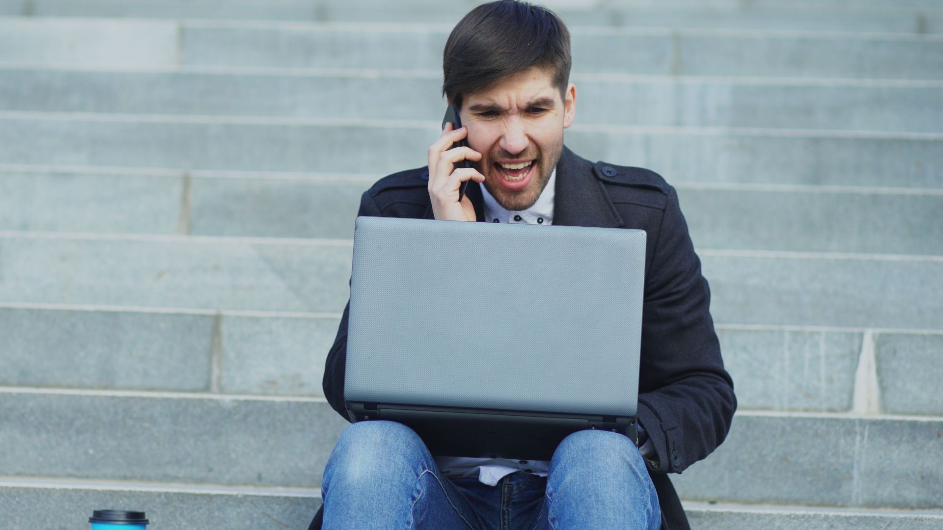 A young man sitting on stairs, using a laptop and talking on the phone, with a coffee cup beside him.